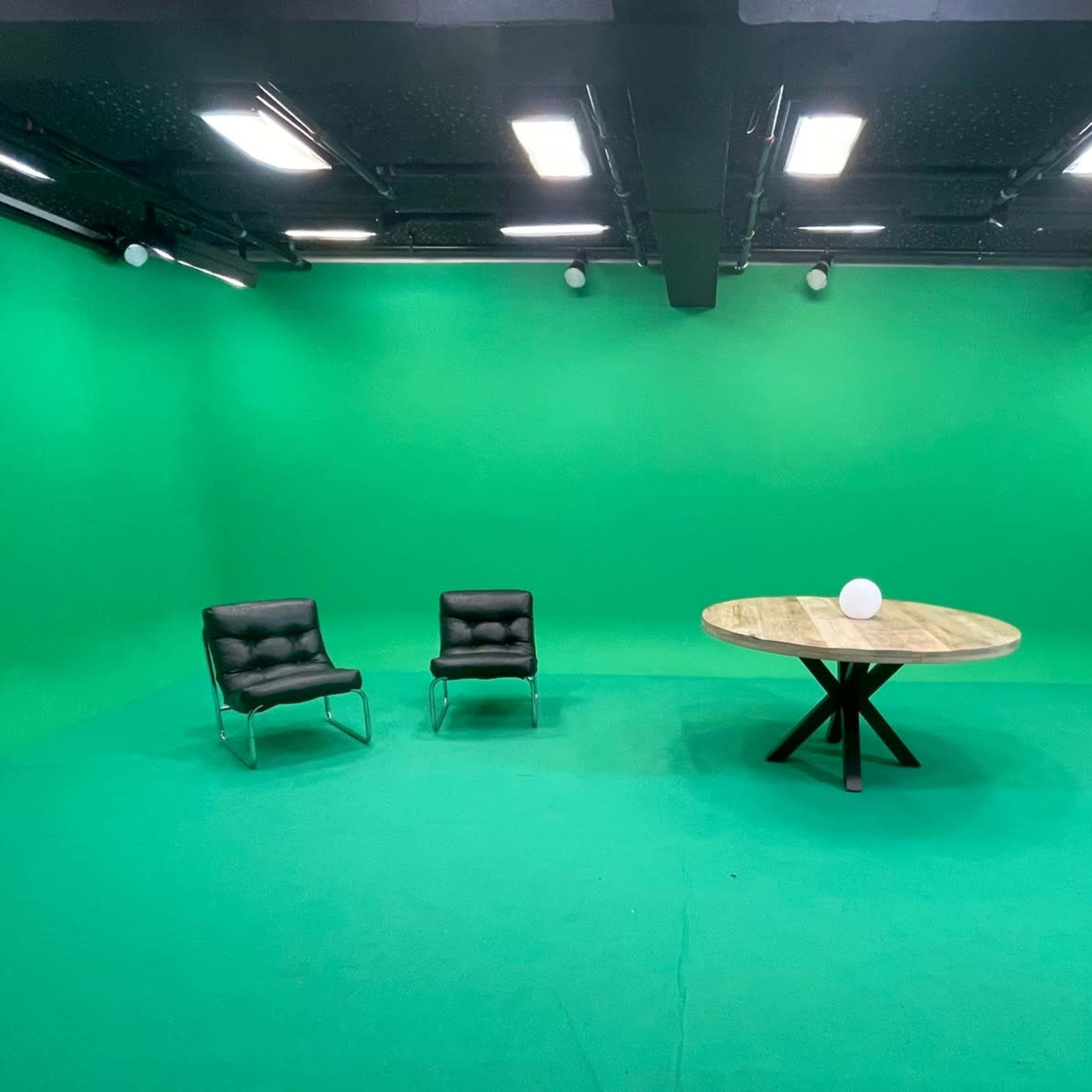 A green-screen studio with two black chairs and a round wooden table in the center.