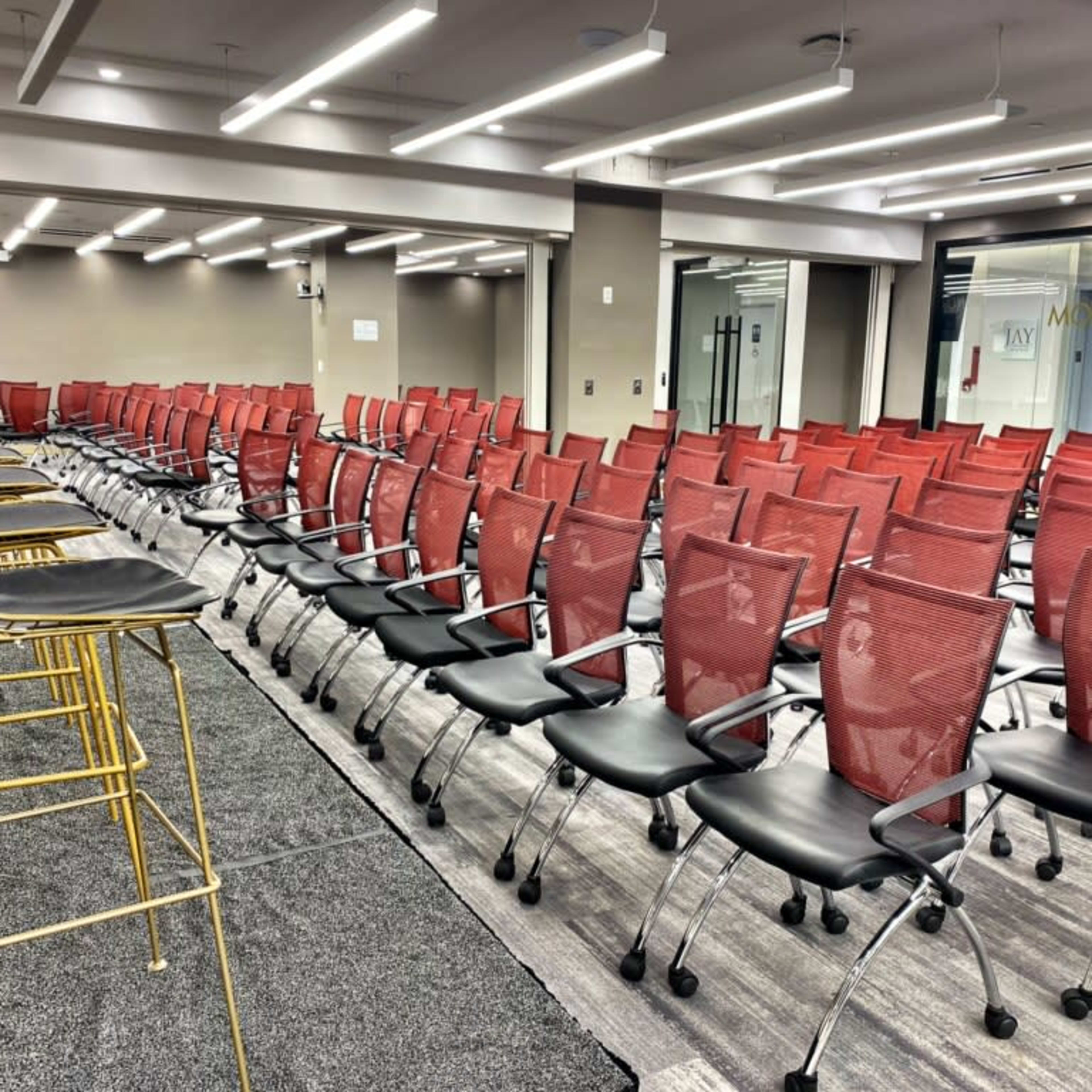A conference room is set up with rows of red mesh chairs and several gold wire stools positioned at the front.