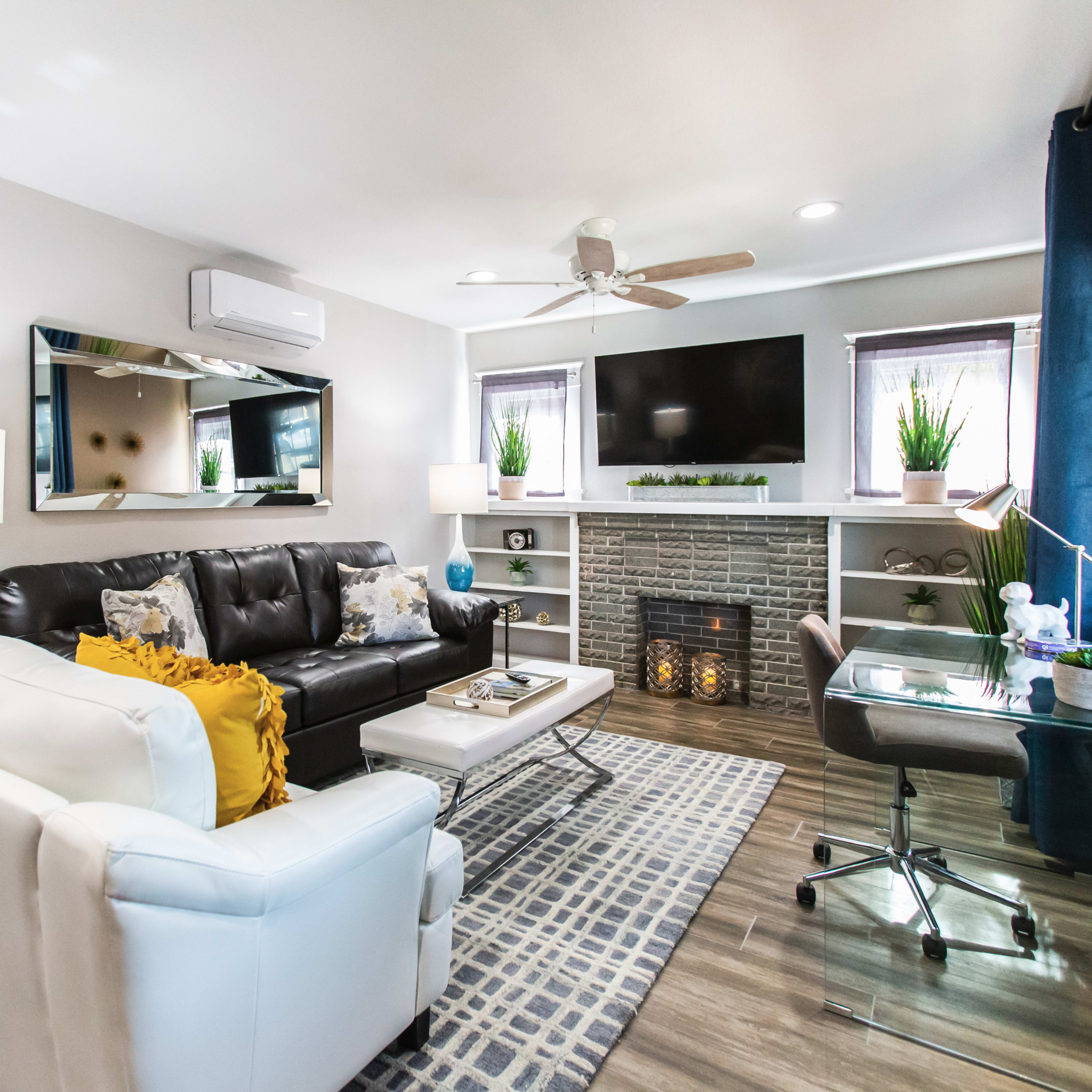 The living room features a black leather sofa, a white armchair, a television mounted above a decorative fireplace, and a desk beside large windows.