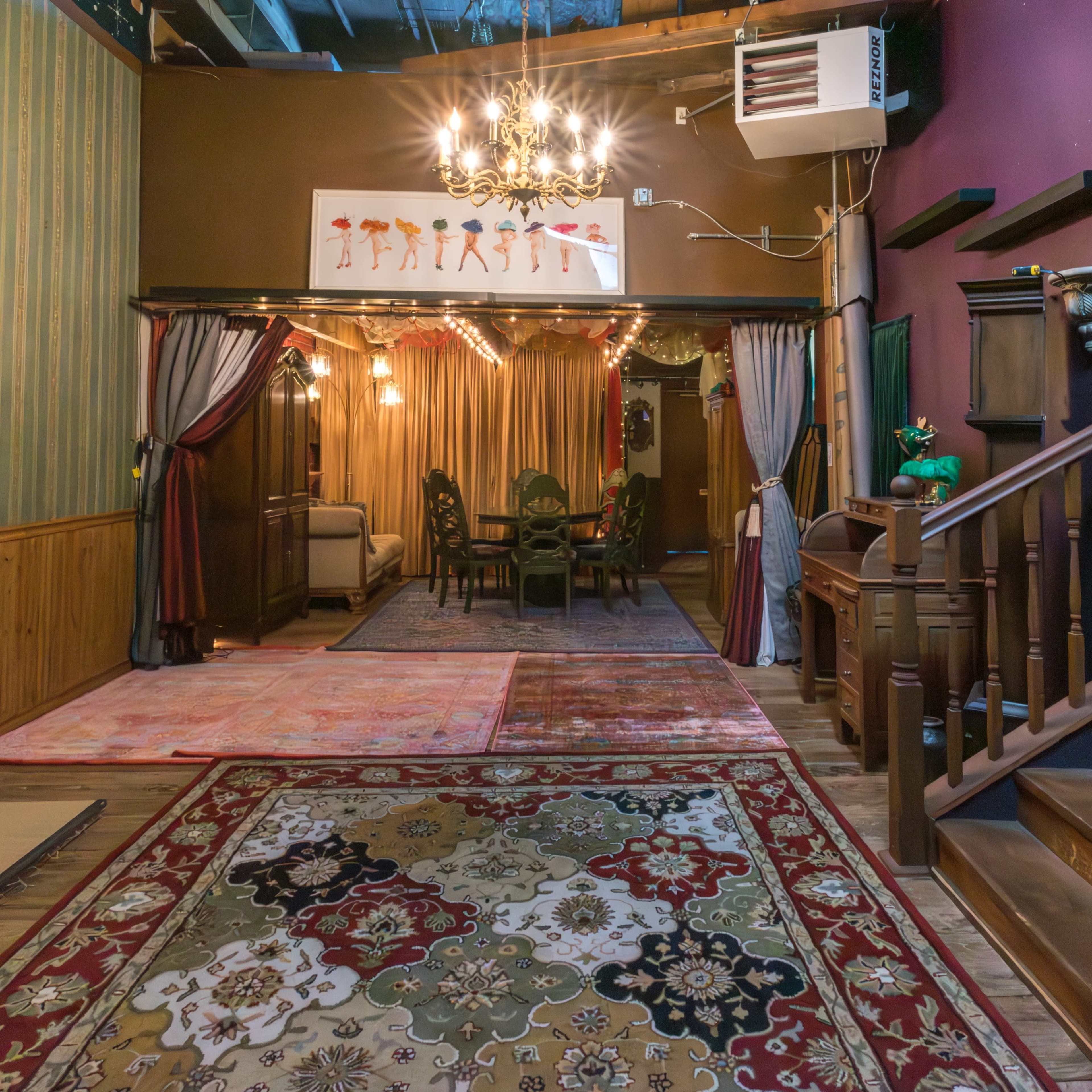 A room with ornate carpets, a chandelier, and a dining area visible through a doorway, featuring patterned curtains and wooden details.