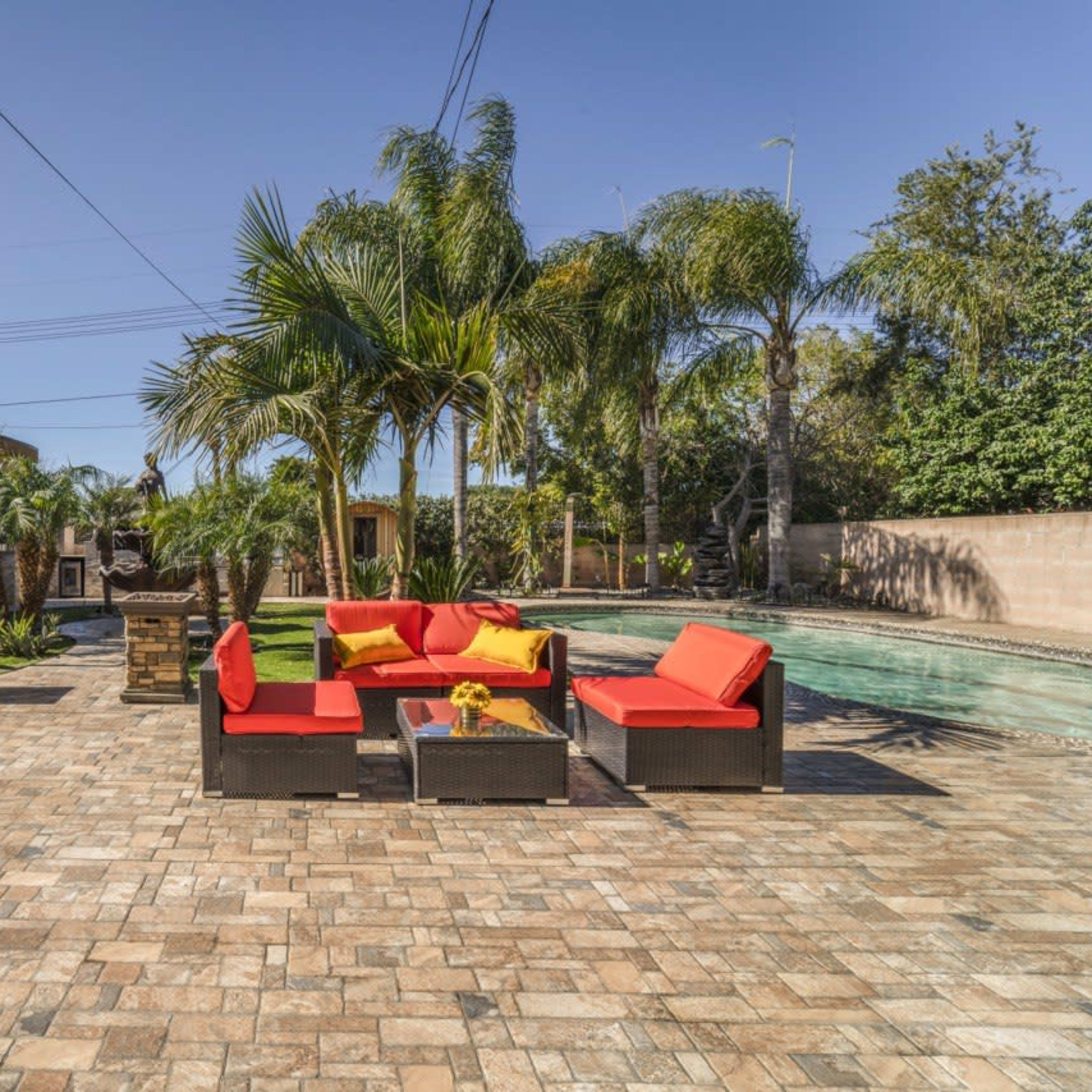 A backyard with a stone patio, a pool, palm trees, and a seating area featuring red and yellow cushions.