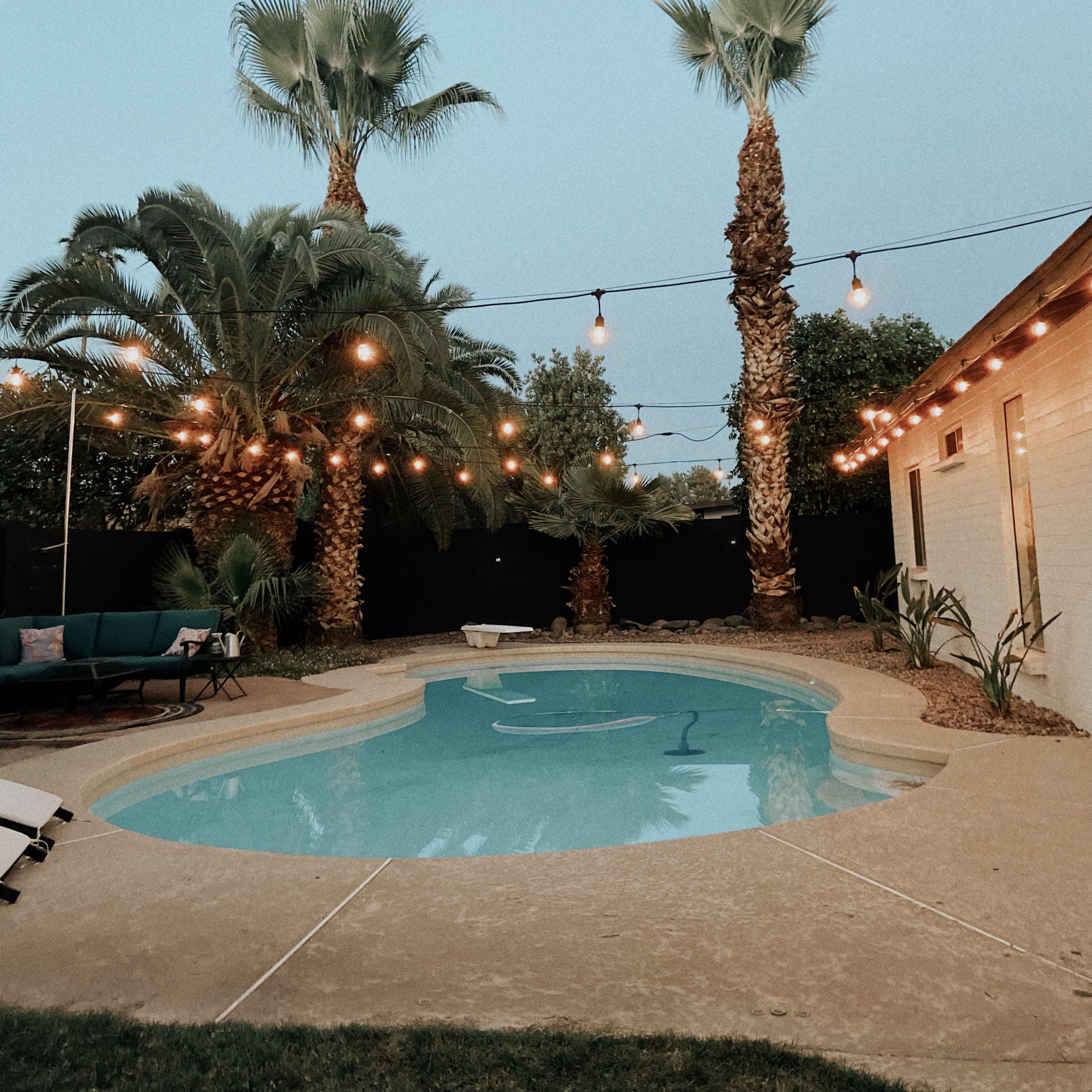 A well-lit backyard pool surrounded by palm trees and string lights at dusk.