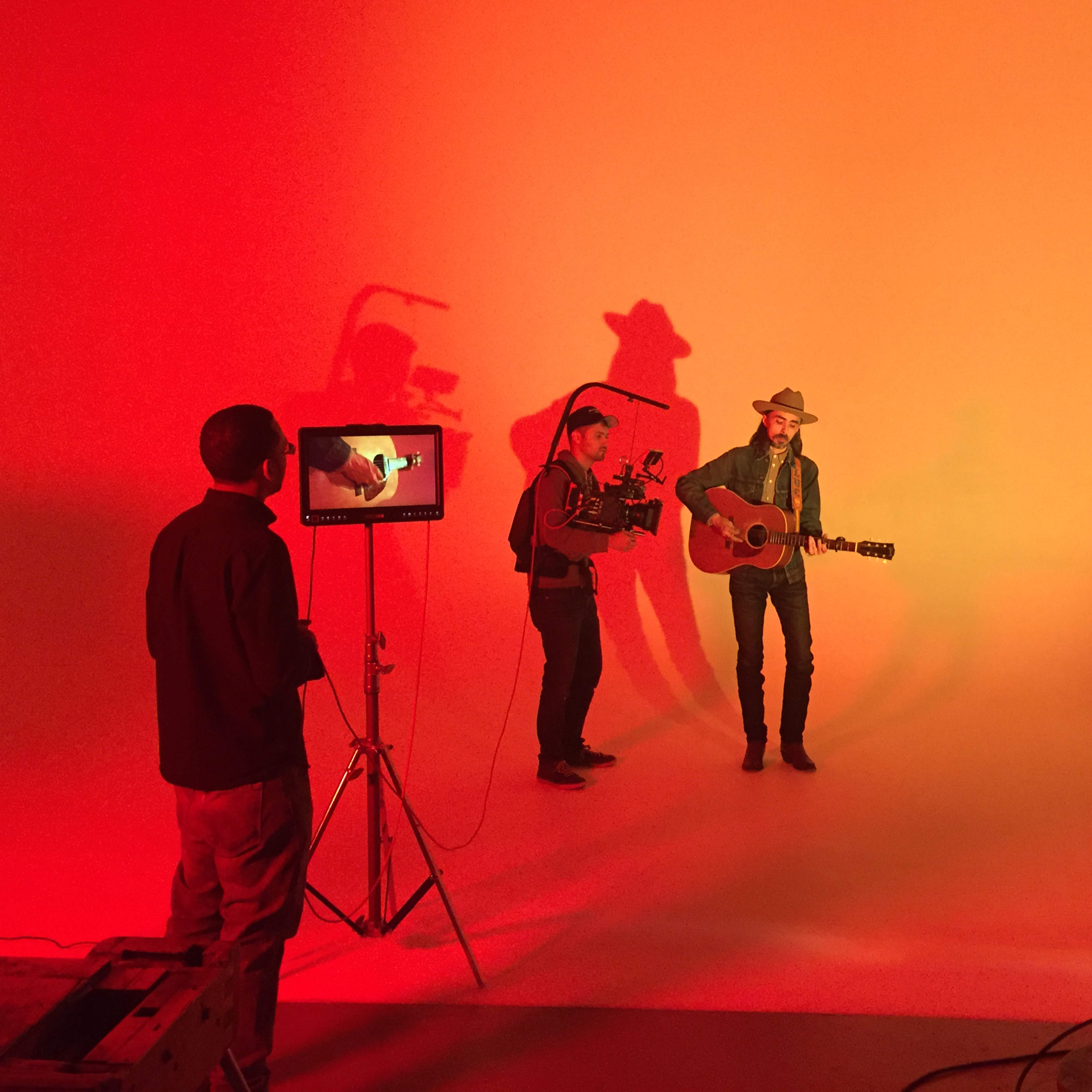 A musician stands with a guitar against a vibrant orange backdrop while two crew members operate cameras and lighting equipment.