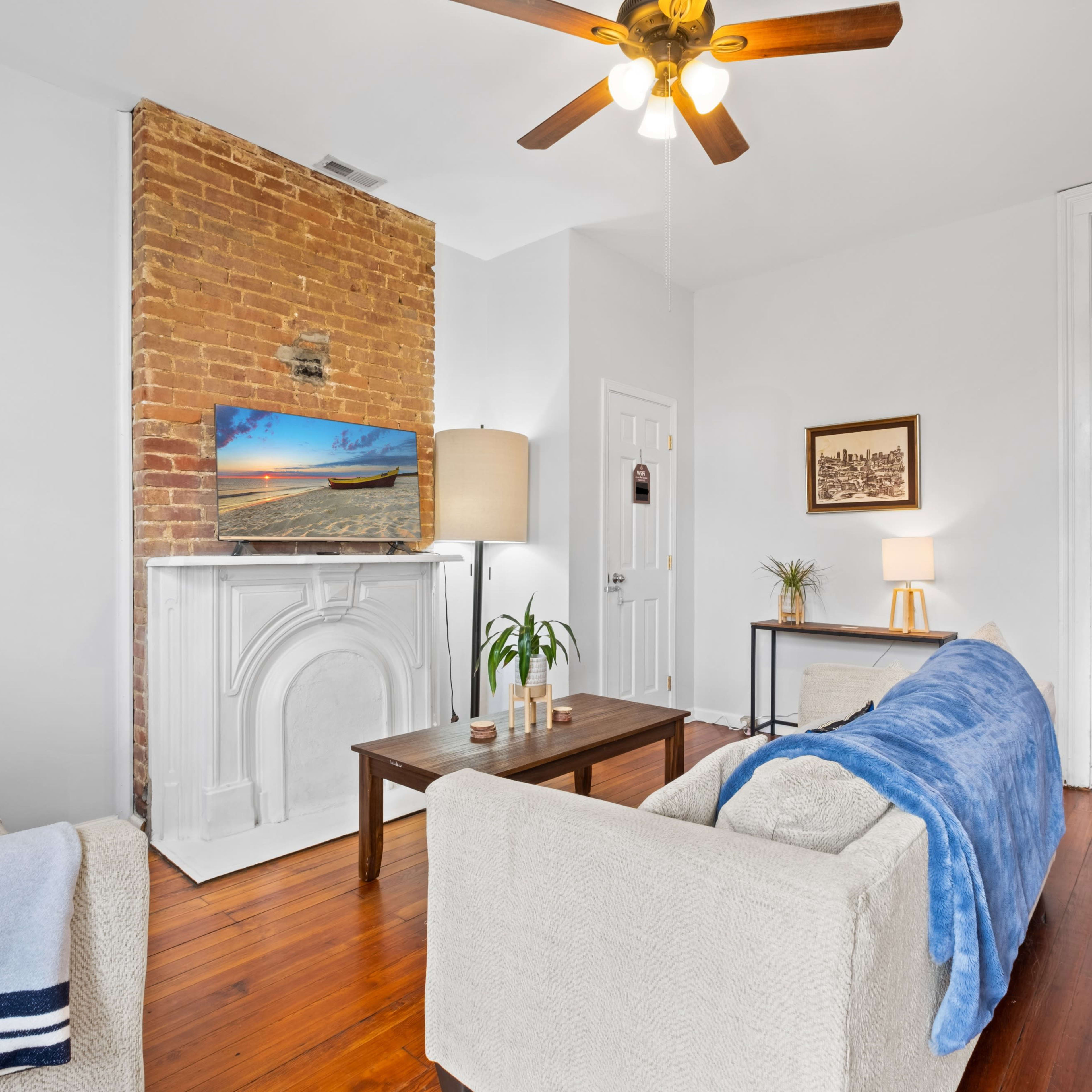The living room features a brick wall, a television mounted above a white mantel, and two sofas with a blue throw blanket on one.