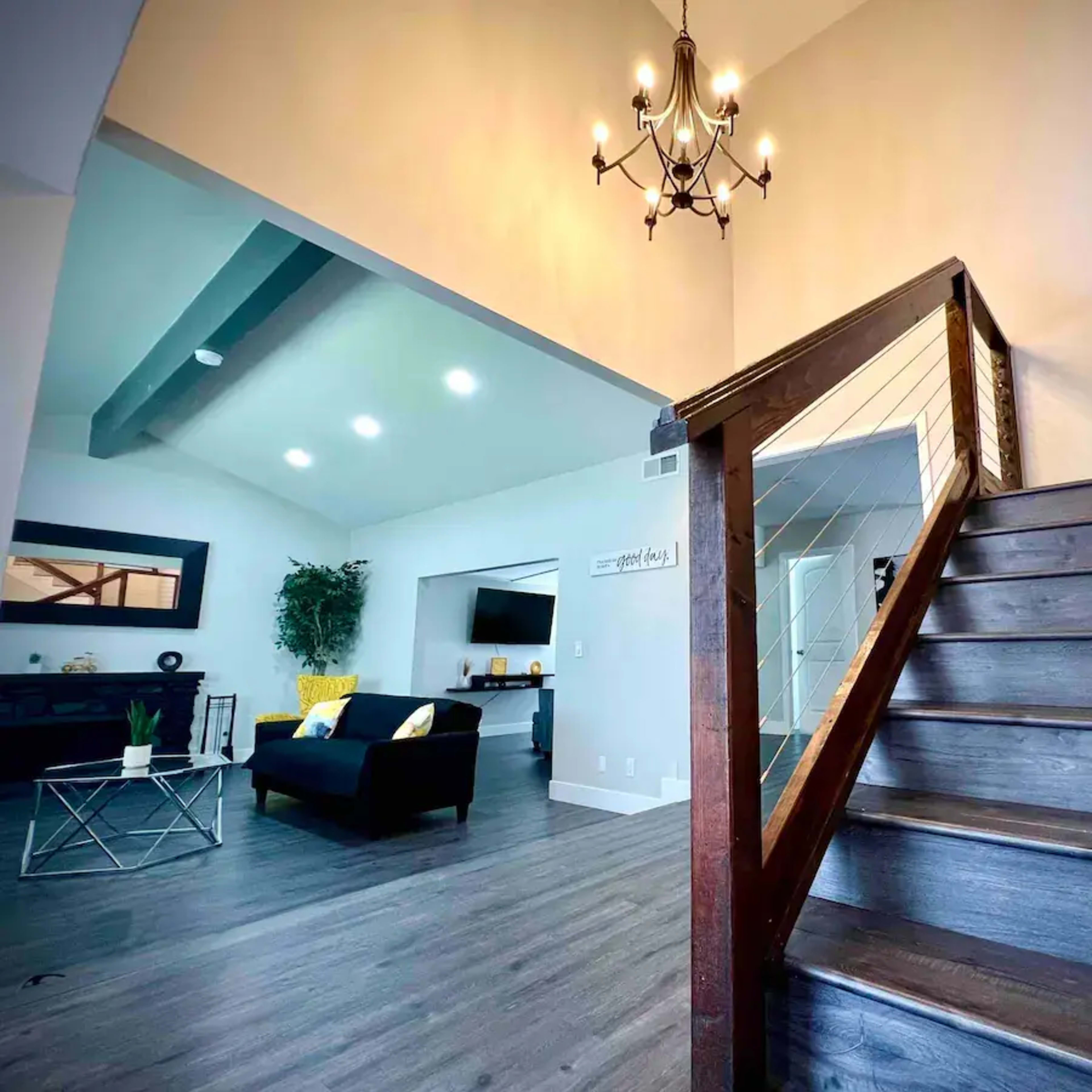 A staircase leads to a bright upper area with a modern chandelier, overlooking a spacious living room furnished with a black sofa and a television.
