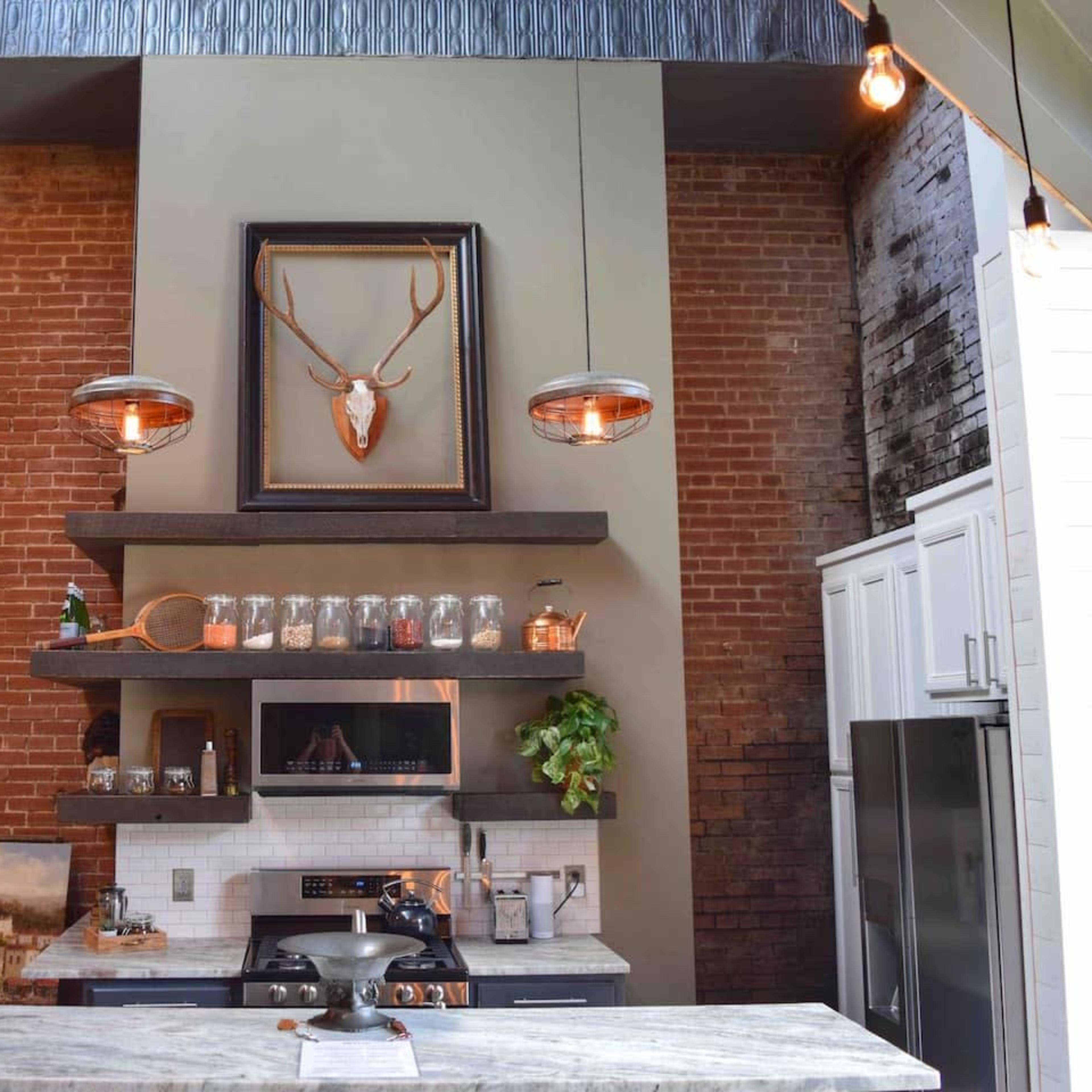 A modern kitchen with exposed brick walls, a prominent shelf displaying jars, and pendant lights hanging above a marble countertop.