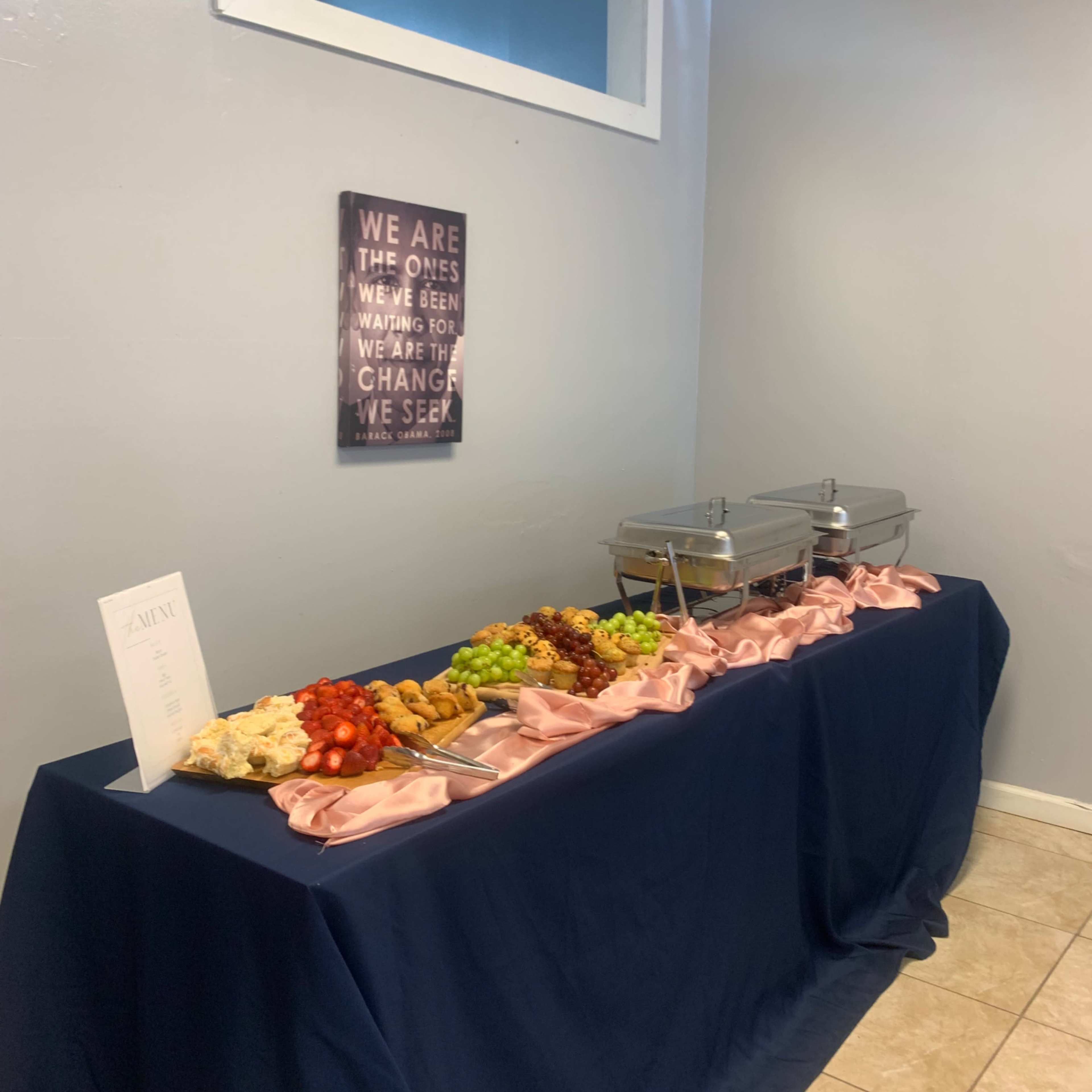 A table covered with a navy blue cloth displays a buffet arrangement of food, including fruit, pastries, and two covered trays.