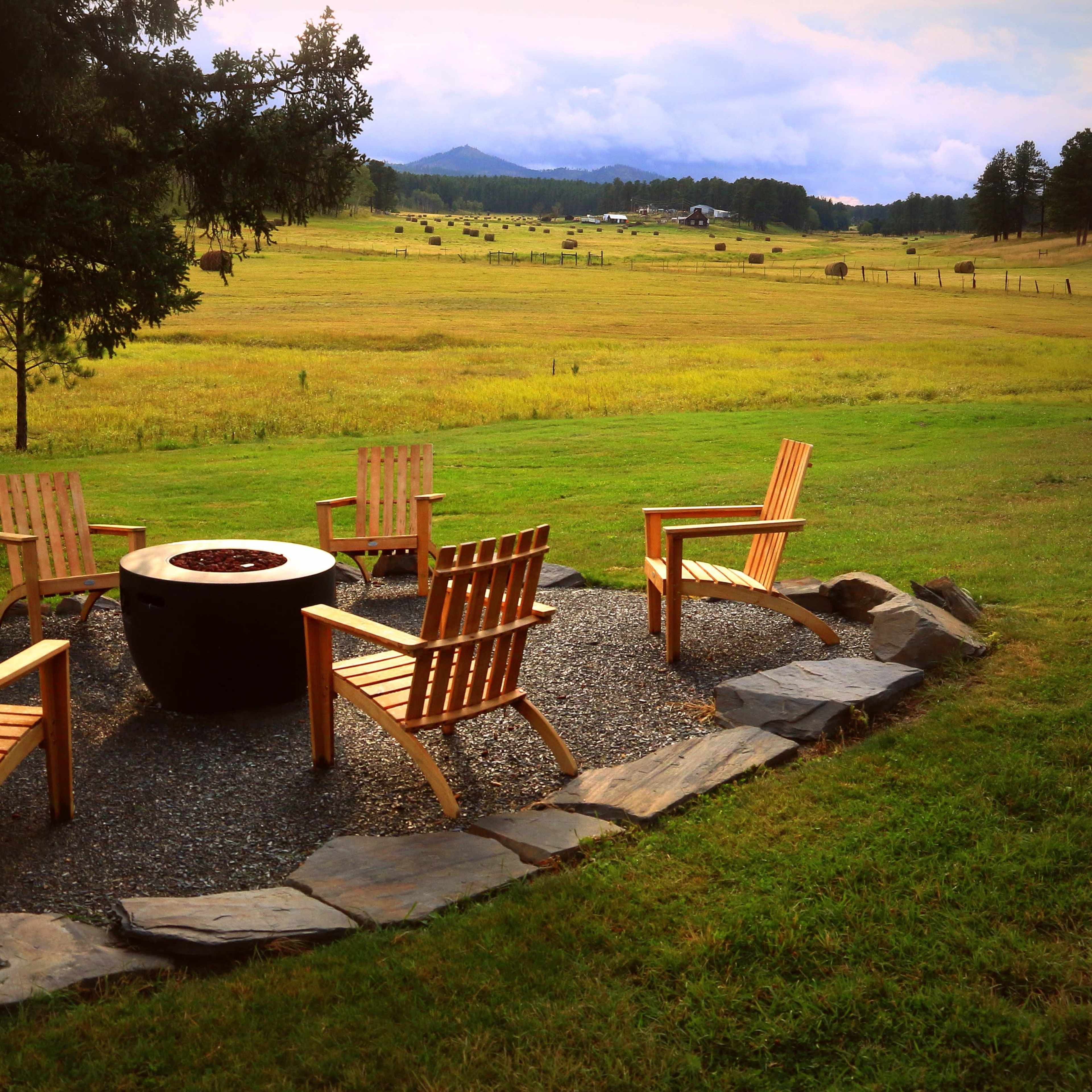 A circular arrangement of wooden chairs surrounds a stone fire pit in a grassy area with a backdrop of rolling hills.