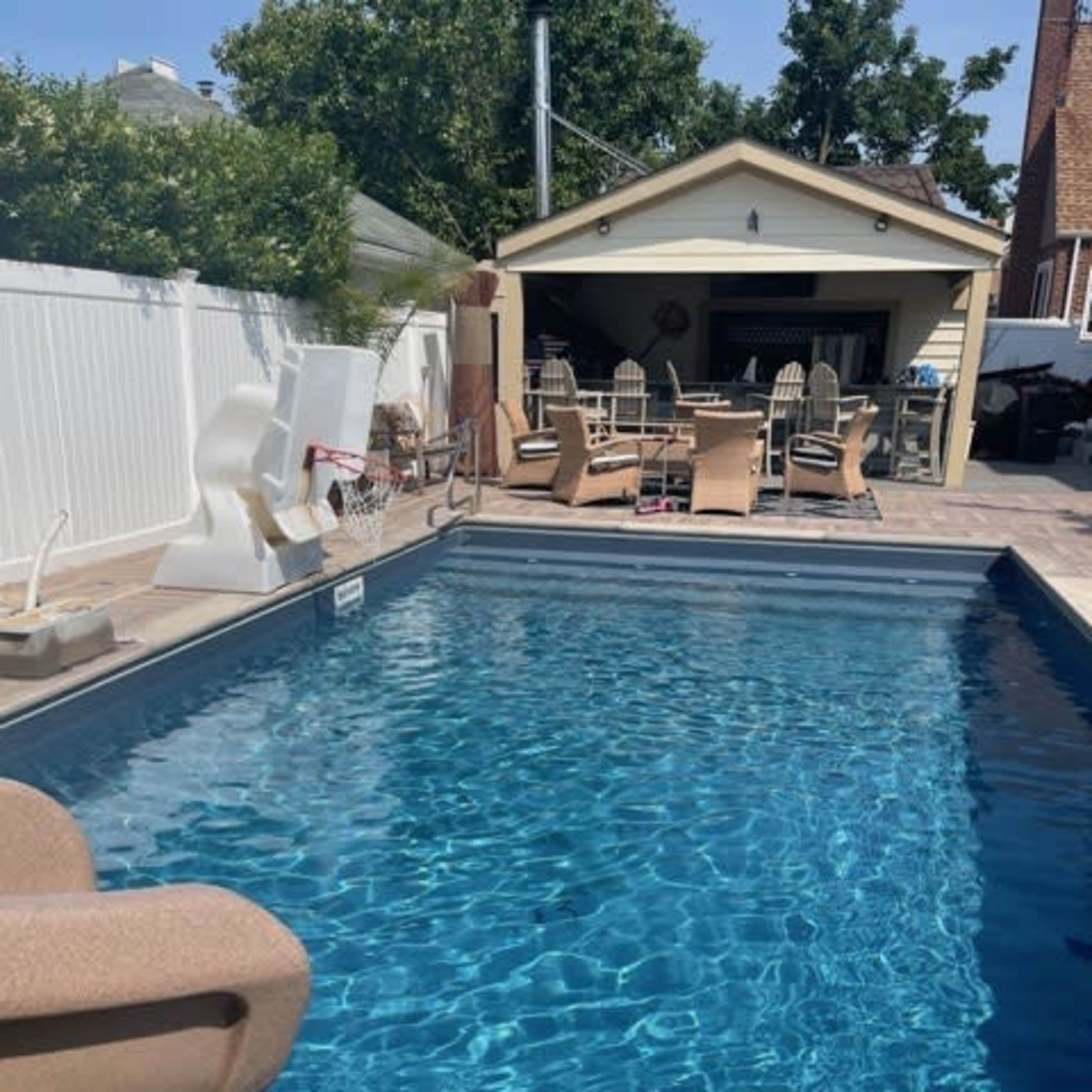 A clear swimming pool with a slide beside a patio area featuring outdoor furniture and a shaded structure.