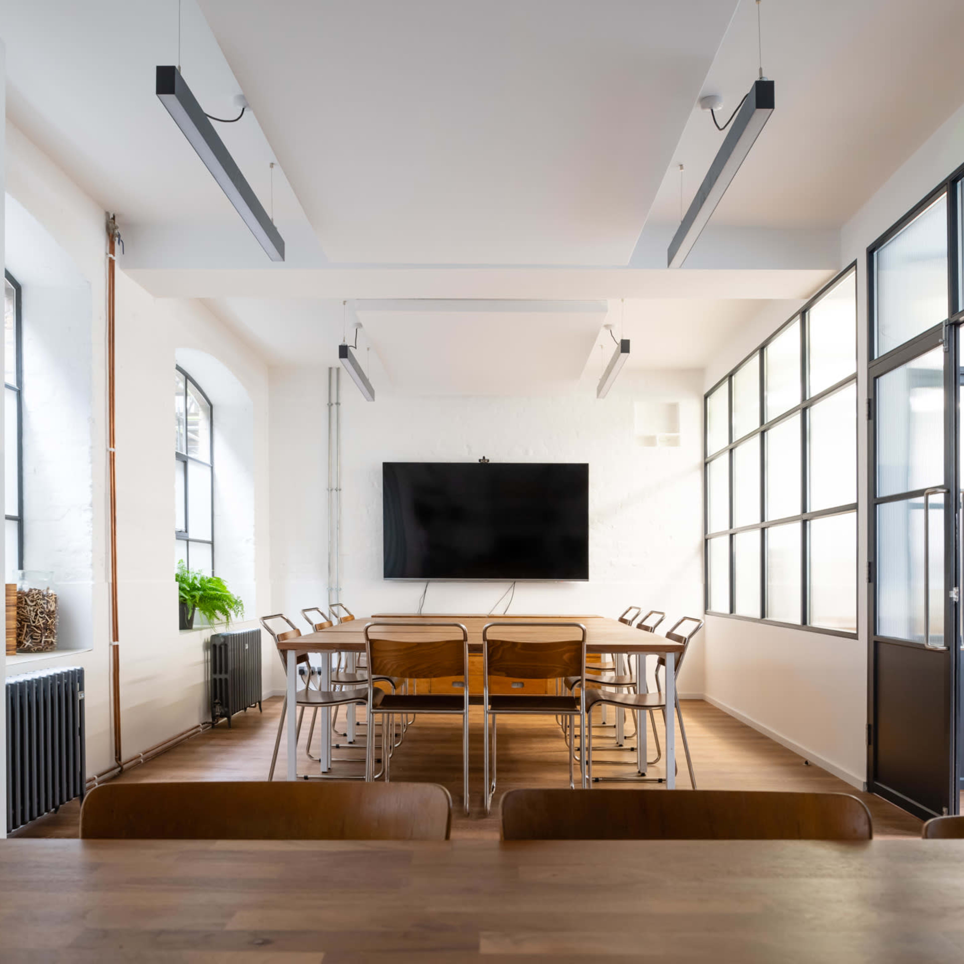 A modern conference room features a long wooden table surrounded by chairs, with a large television mounted on the wall and large windows providing natural light.