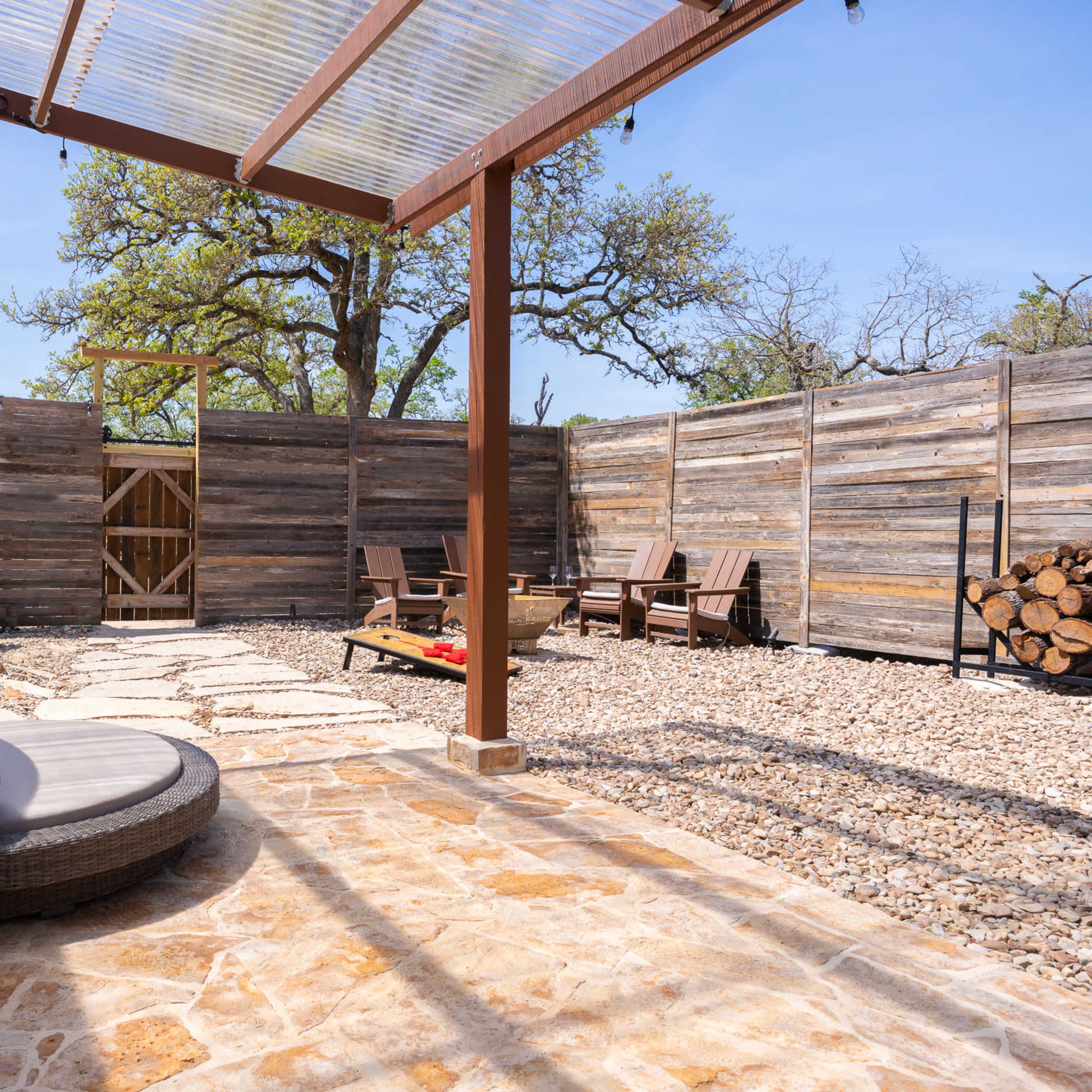 A spacious outdoor patio area featuring a circular lounge chair, stone flooring, wooden fence, and minimal seating arrangements against a backdrop of trees.
