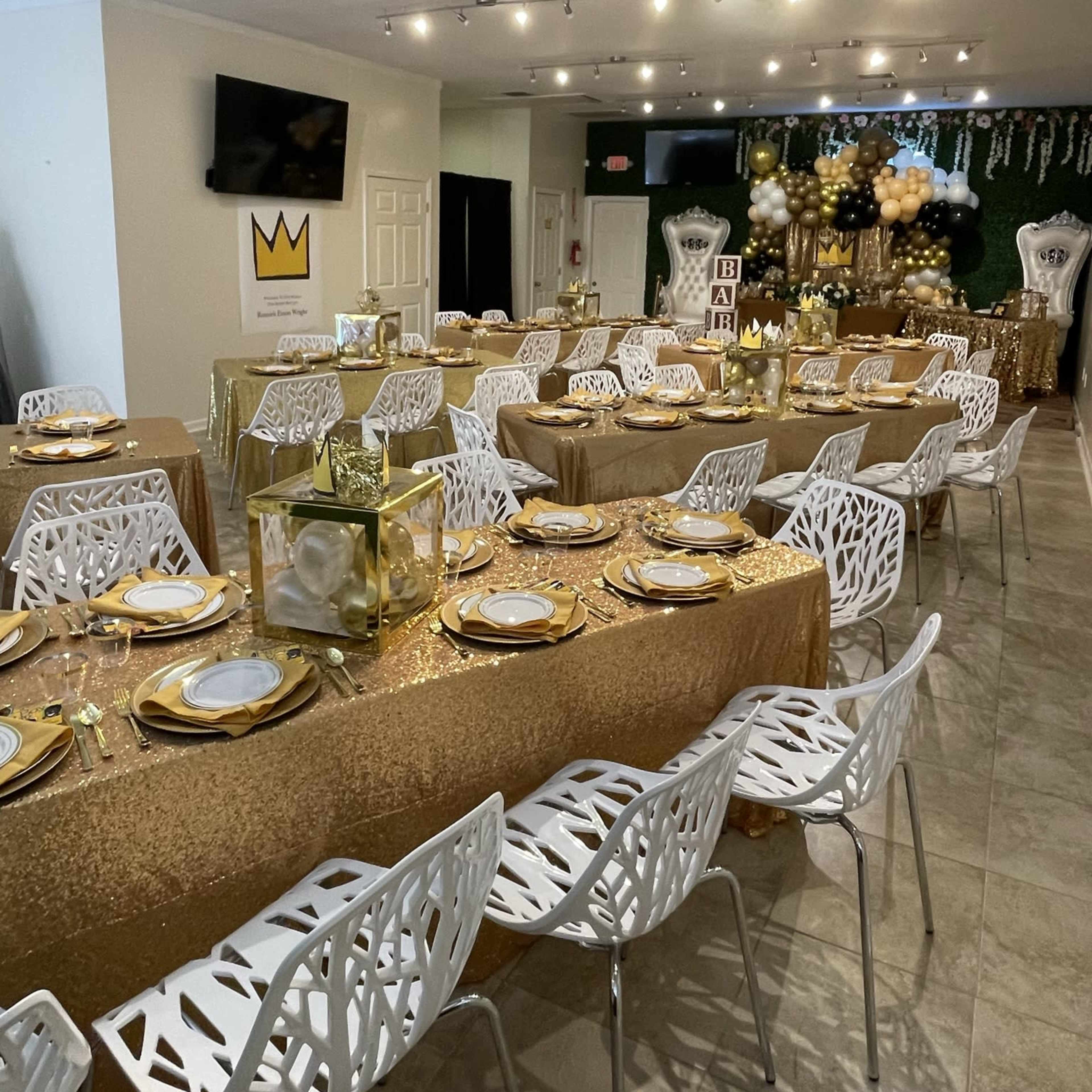 A decorated event space with multiple tables covered in gold tablecloths and white chairs, adorned with elegant table settings and decorative centerpieces.