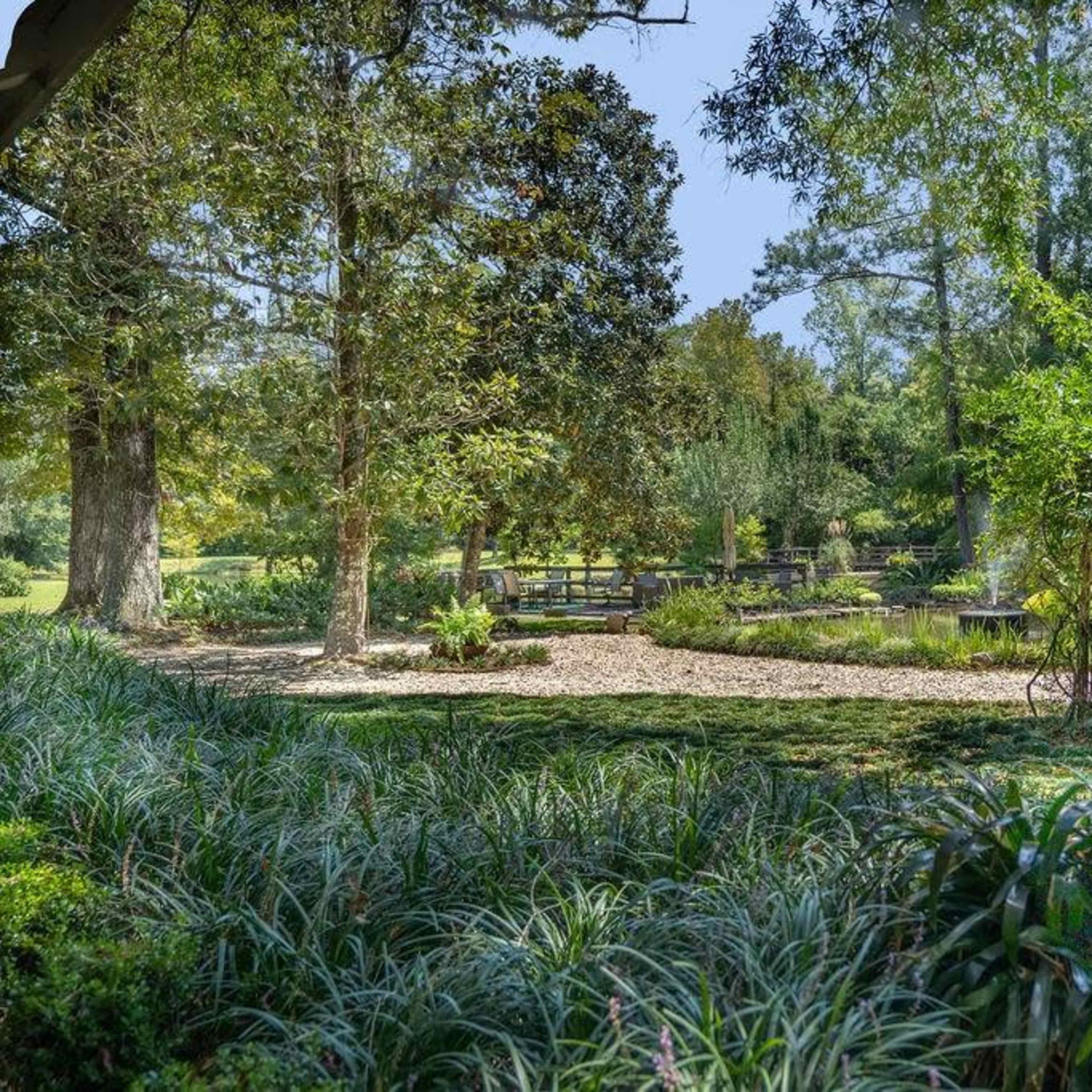 A garden with a gravel path surrounded by lush greenery and trees, featuring a small pond and wooden benches in the background.