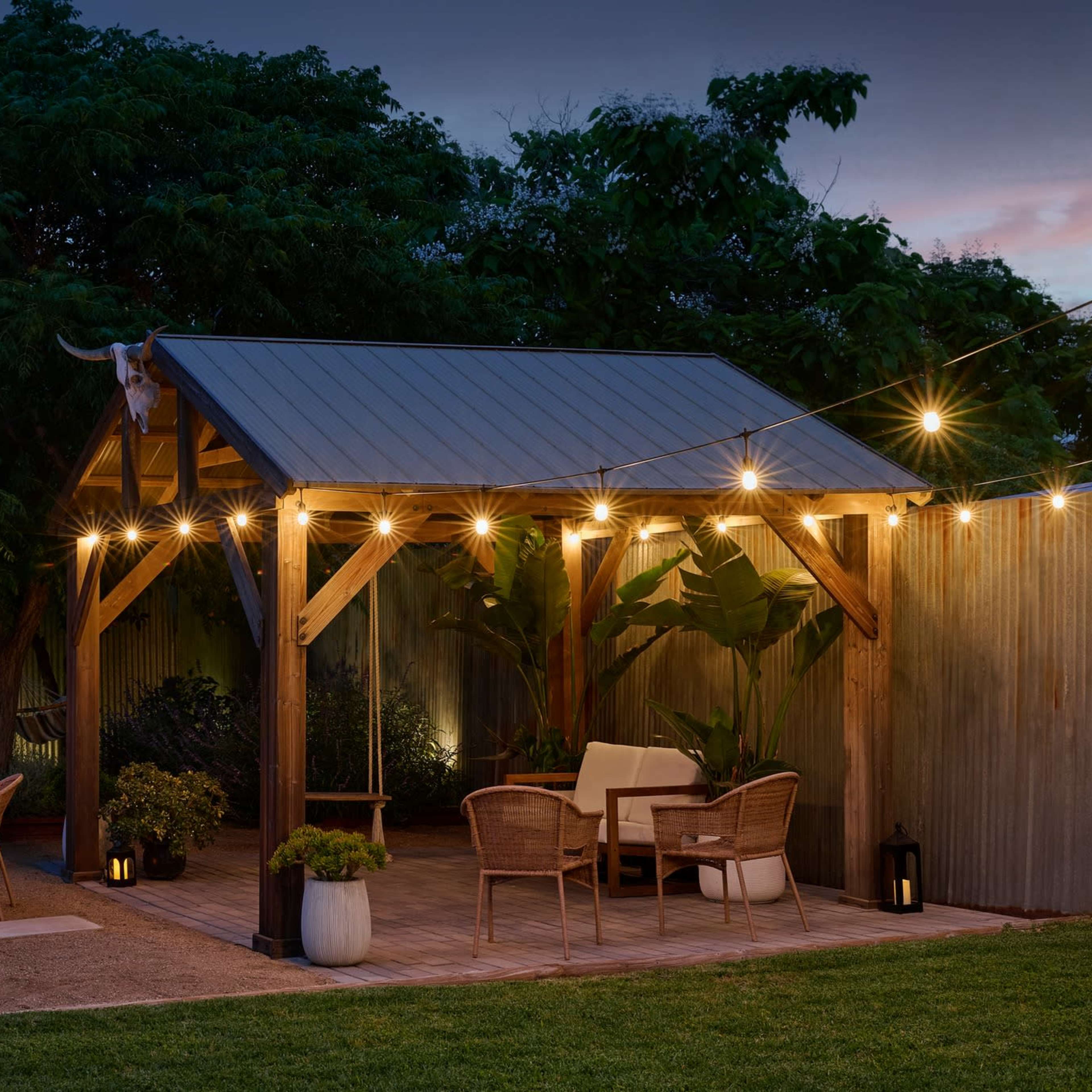 A wooden pavilion with string lights is set up in a landscaped backyard, featuring seating areas around a fire pit.