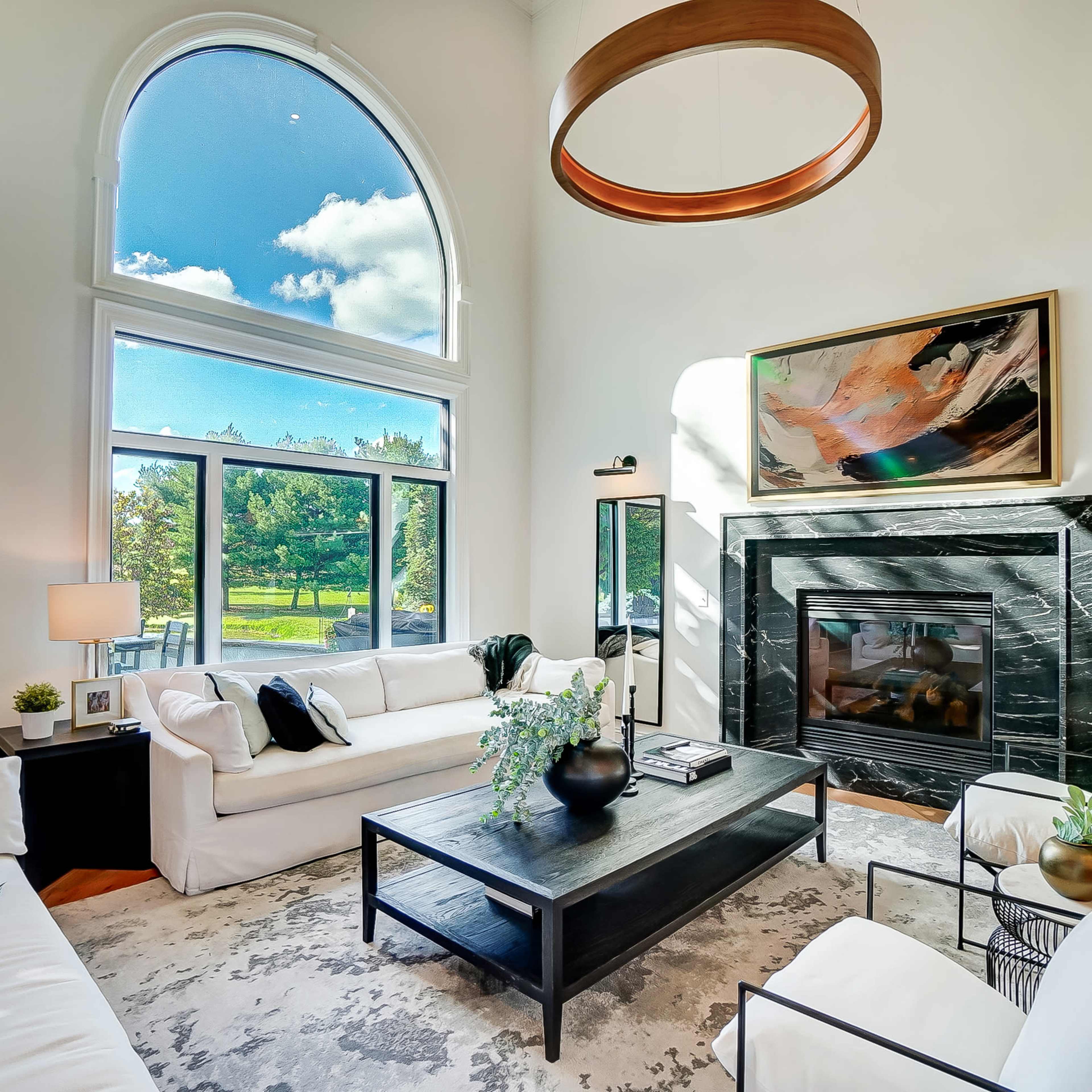 A bright living room features large windows overlooking greenery, a black stone fireplace, and modern furnishings.