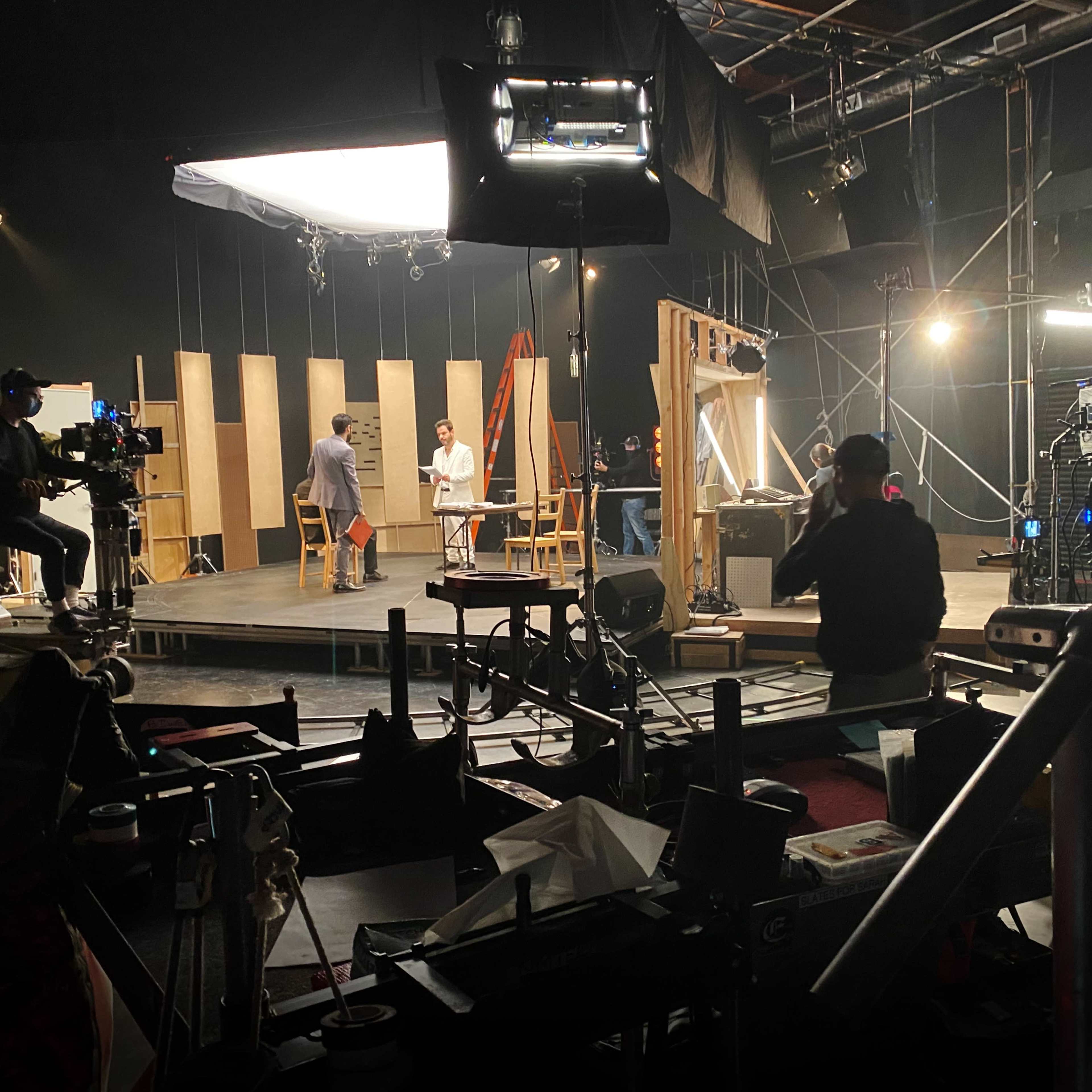 A film or television production set features crew members and equipment arranged around a central stage with lighting and backdrop elements.