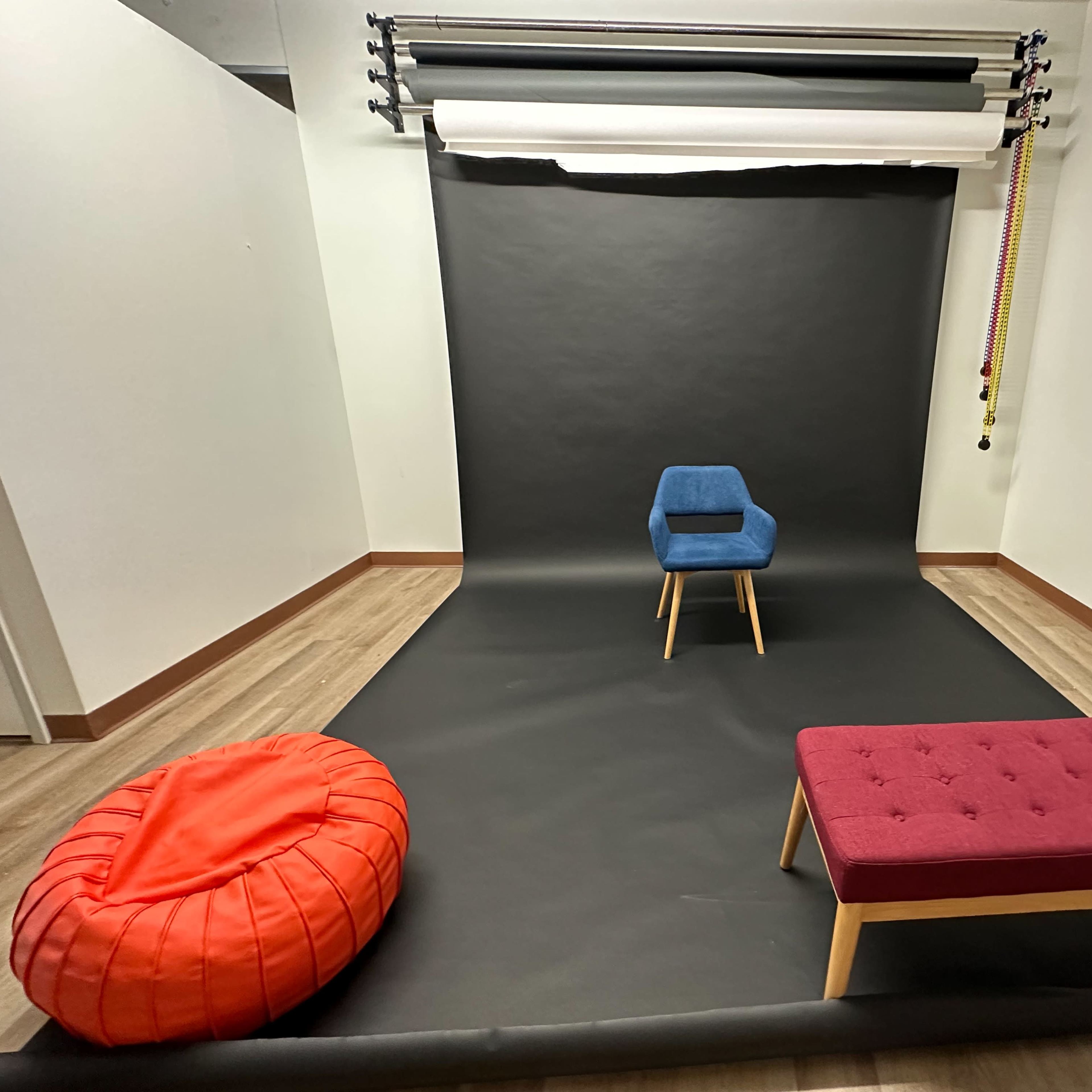 A minimalist studio setup with a black backdrop, a blue chair on a wooden frame, a red bean bag, and a purple bench.