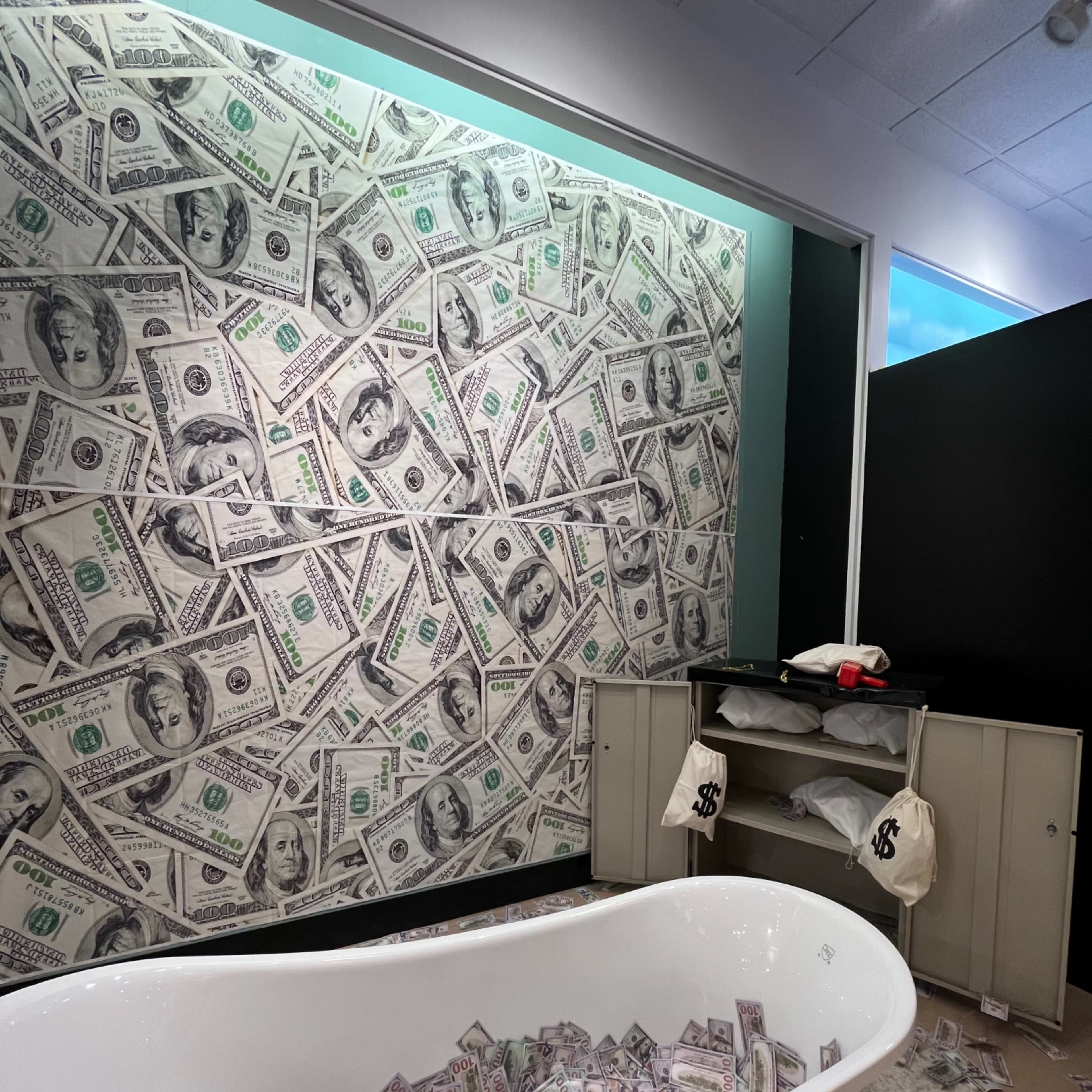 A white bathtub filled with what appears to be fake money sits in a room with a wall covered in a dollar bill design.