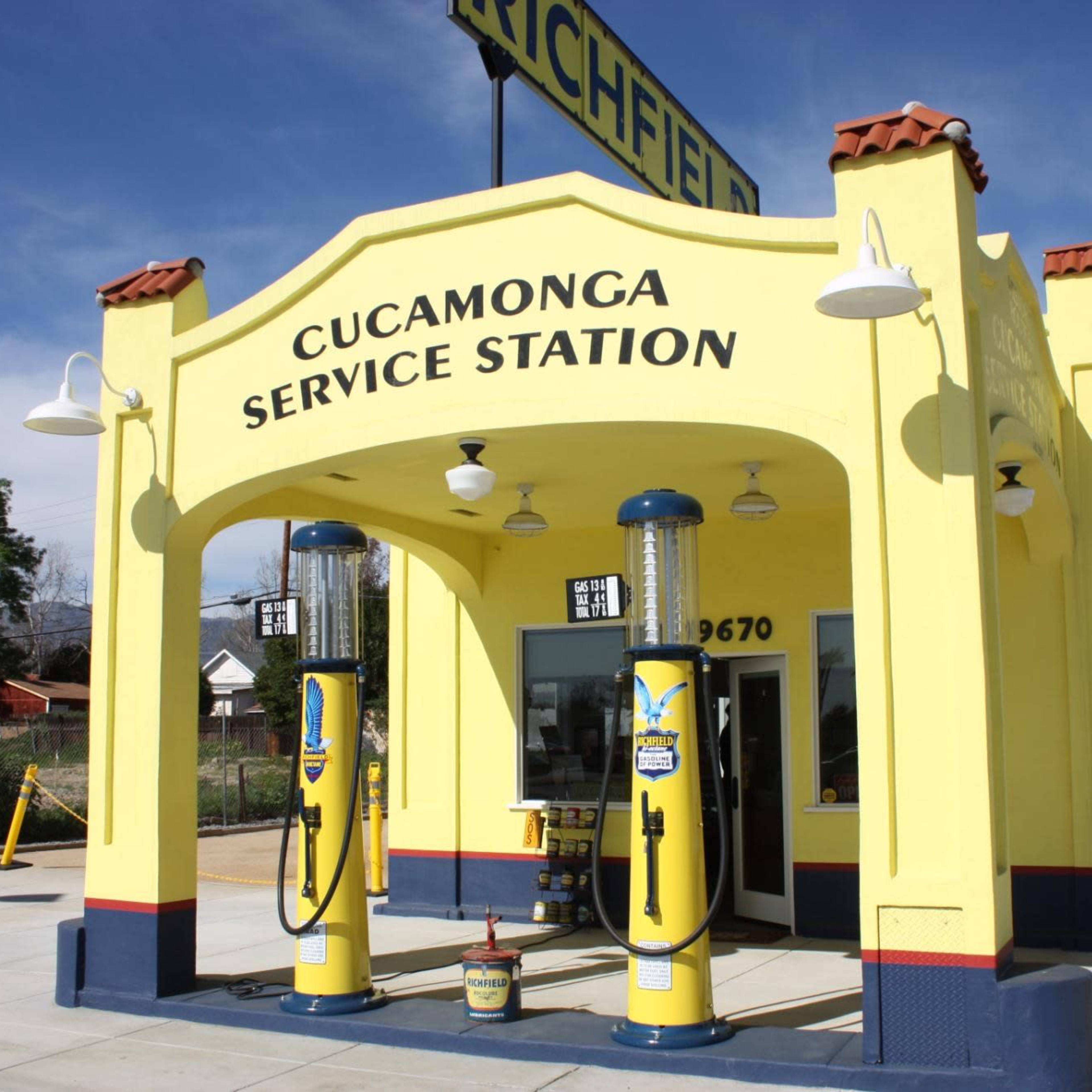 A bright yellow service station featuring vintage gas pumps is situated at the corner of a street with mountains in the background.