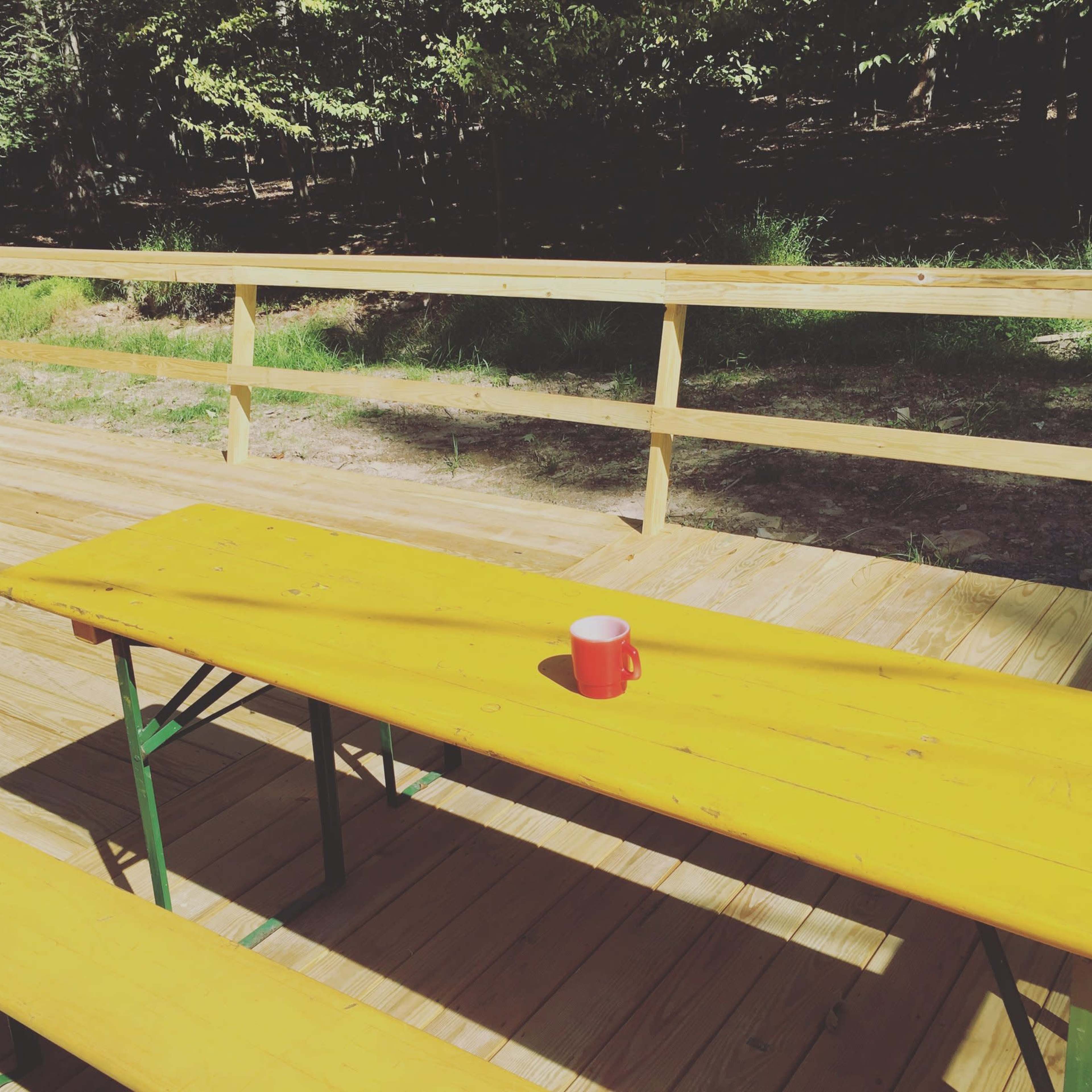 A red mug sits on a yellow wooden table in an outdoor setting surrounded by trees.