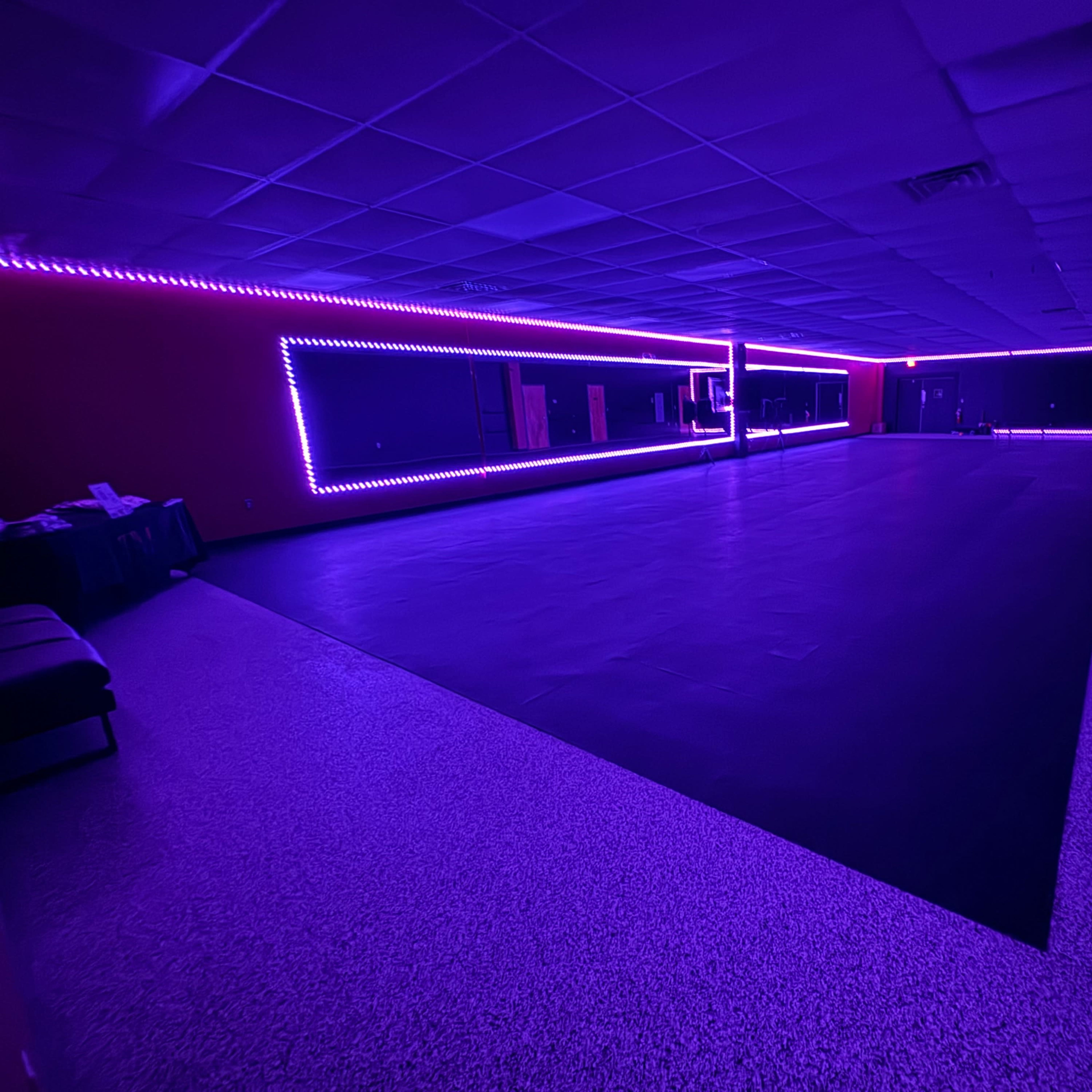 A dimly lit dance studio with purple LED lights highlighting the walls and mirrors.