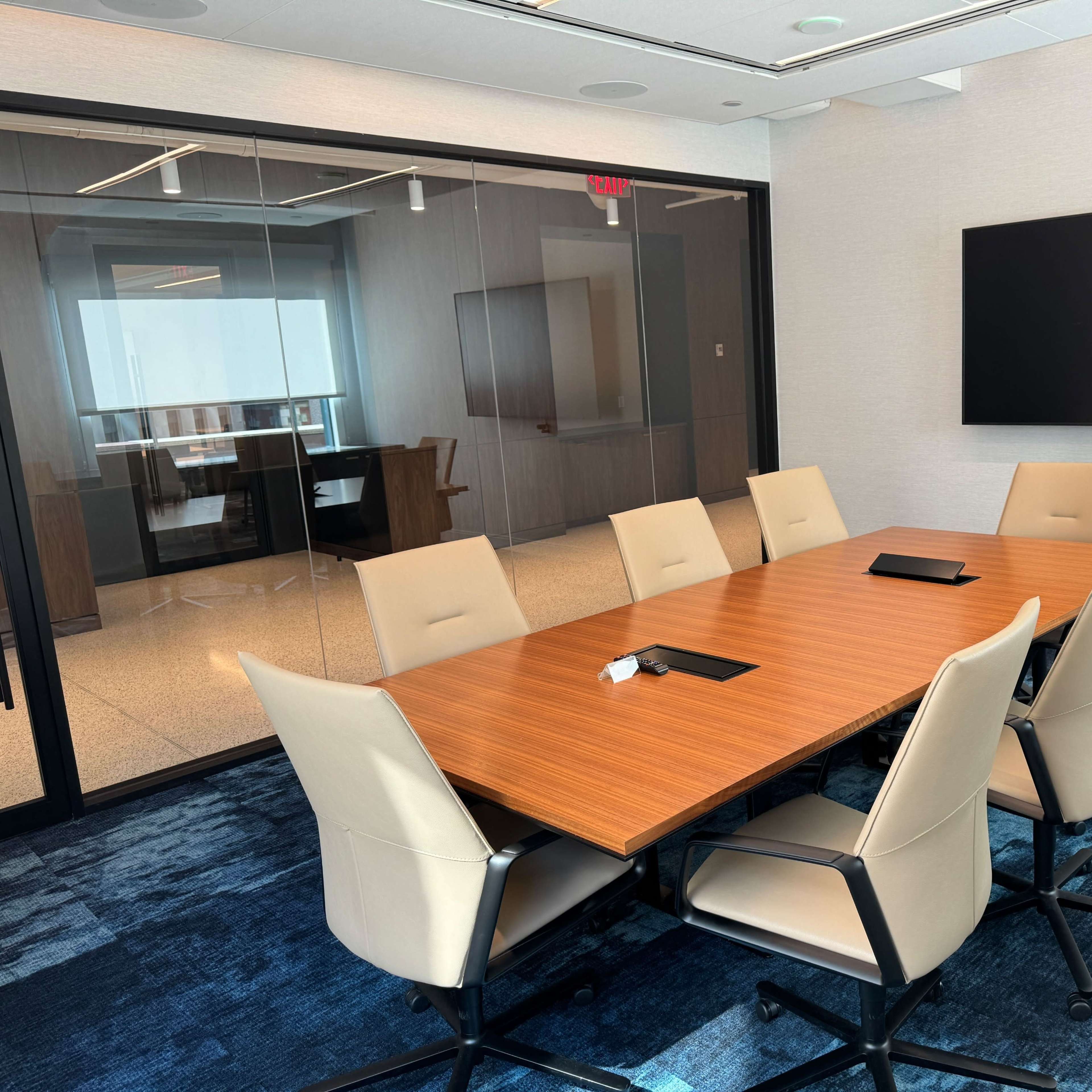 A modern conference room with a long wooden table surrounded by beige chairs, and a large screen mounted on the wall.
