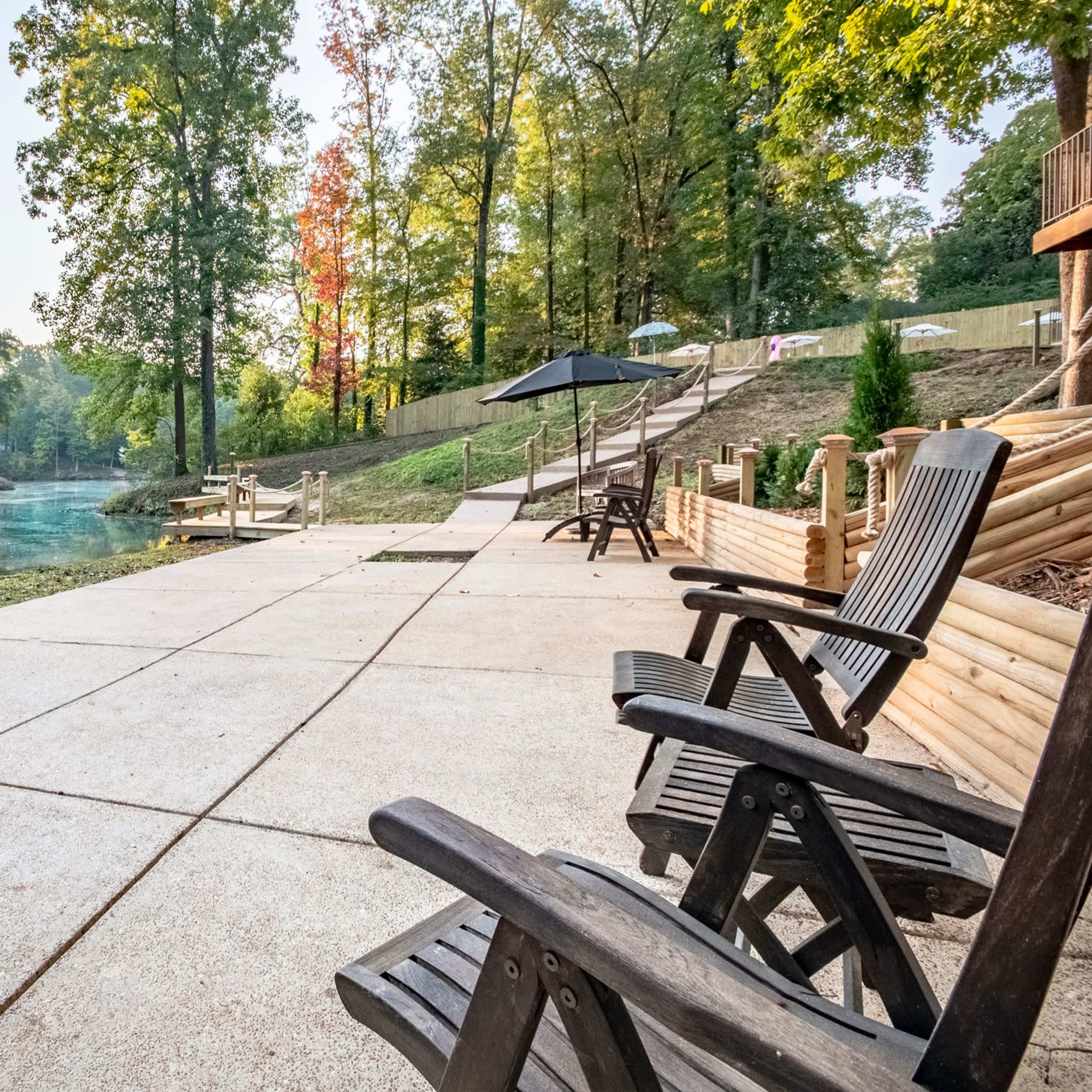 A tranquil riverside patio with wooden chairs and an umbrella, surrounded by trees and a clear waterway.