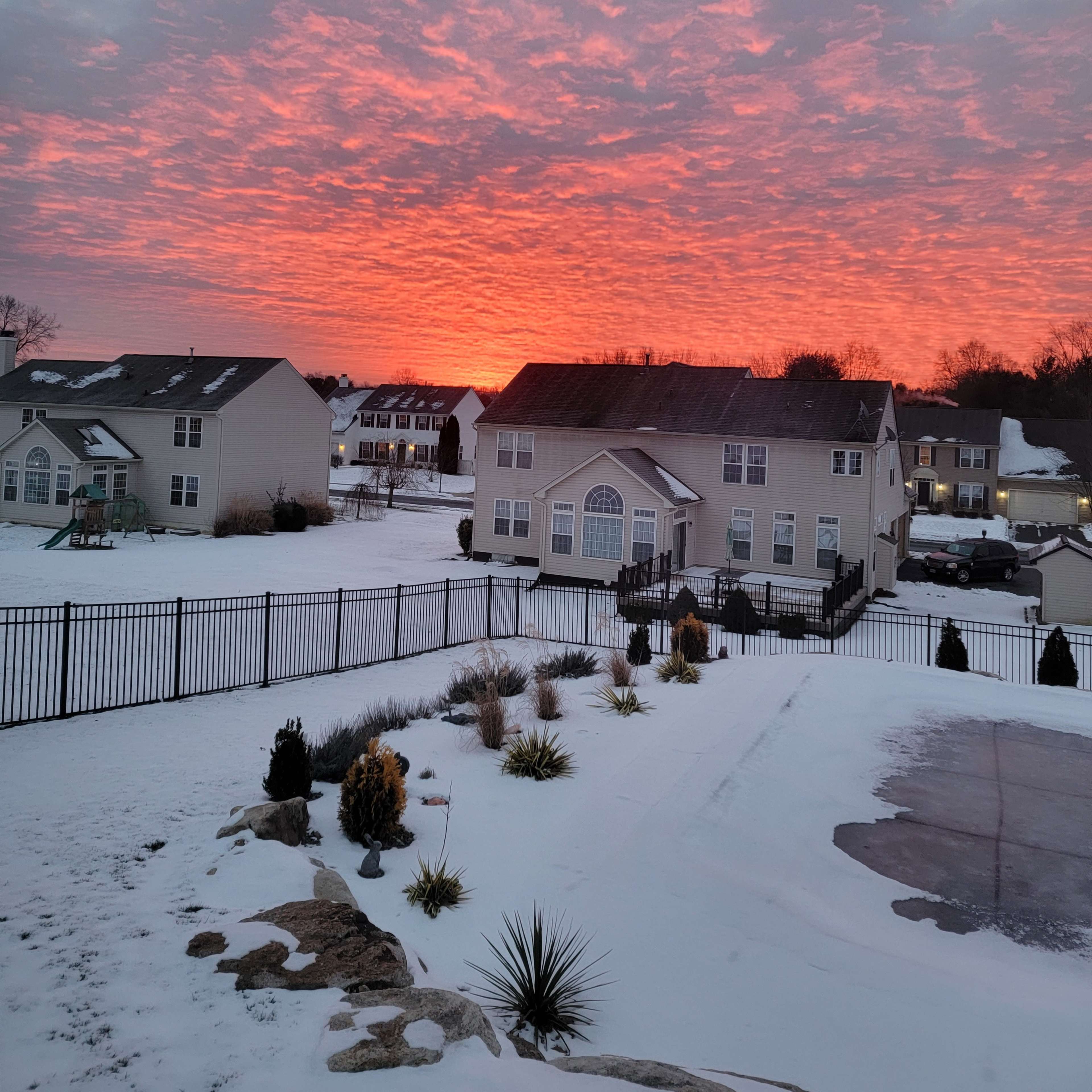 A winter landscape features snow-covered ground with houses in the background and a vibrant sunset illuminating the sky.
