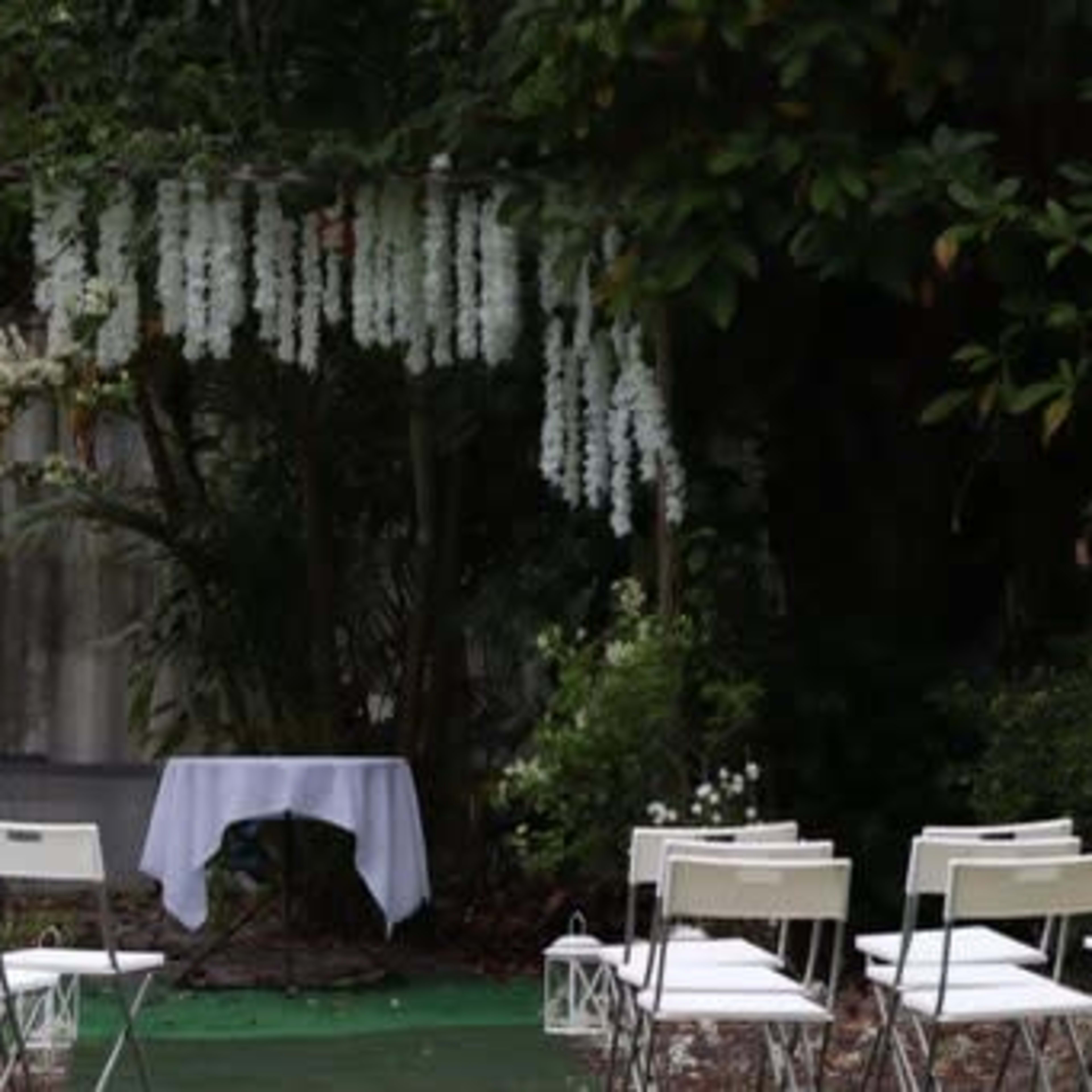 A small outdoor space is set up for a ceremony, featuring white chairs arranged in rows facing a table draped with a white cloth, surrounded by greenery and hanging white flowers.