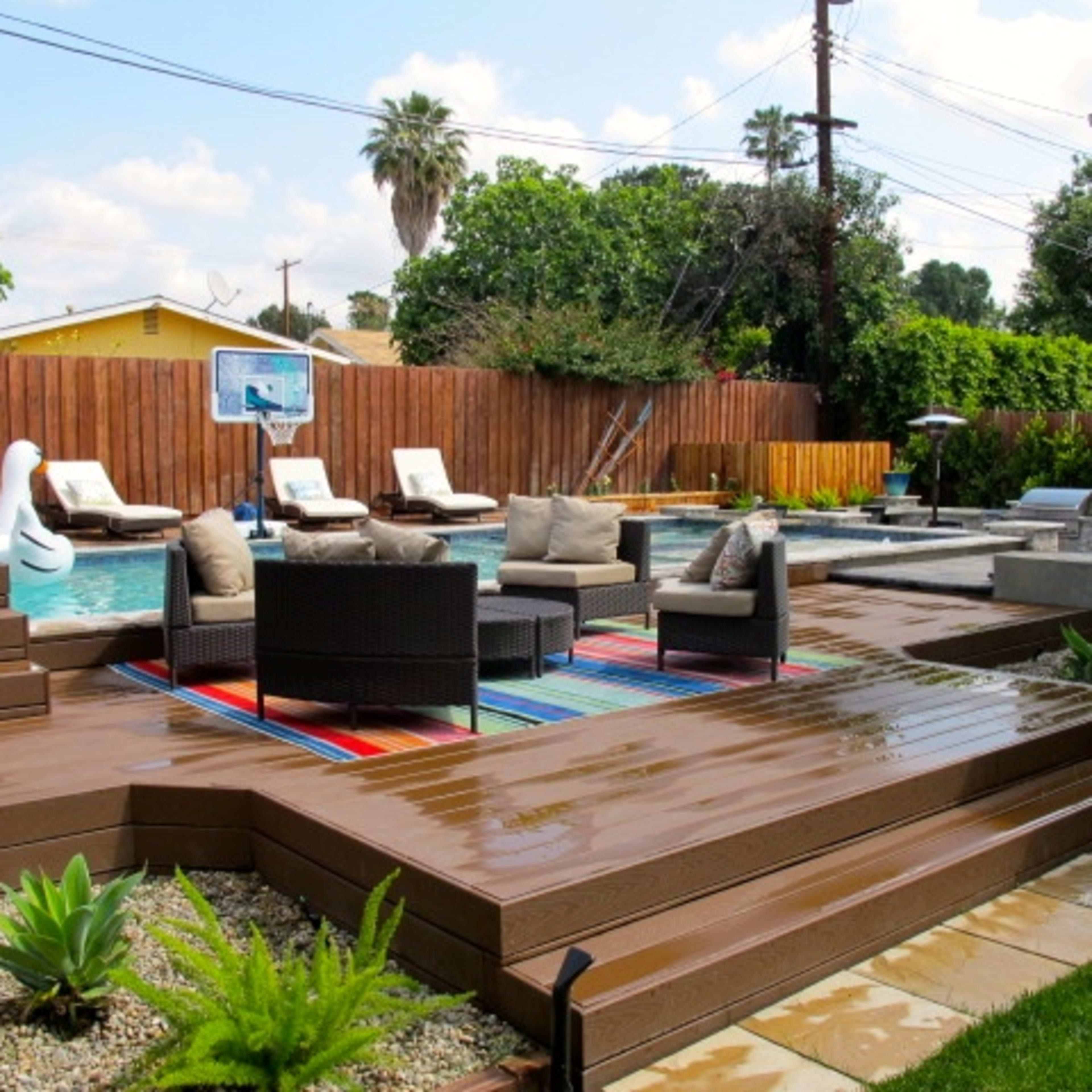 A residential backyard featuring a swimming pool surrounded by lounge chairs, a colorful rug, and landscaped areas with plants.