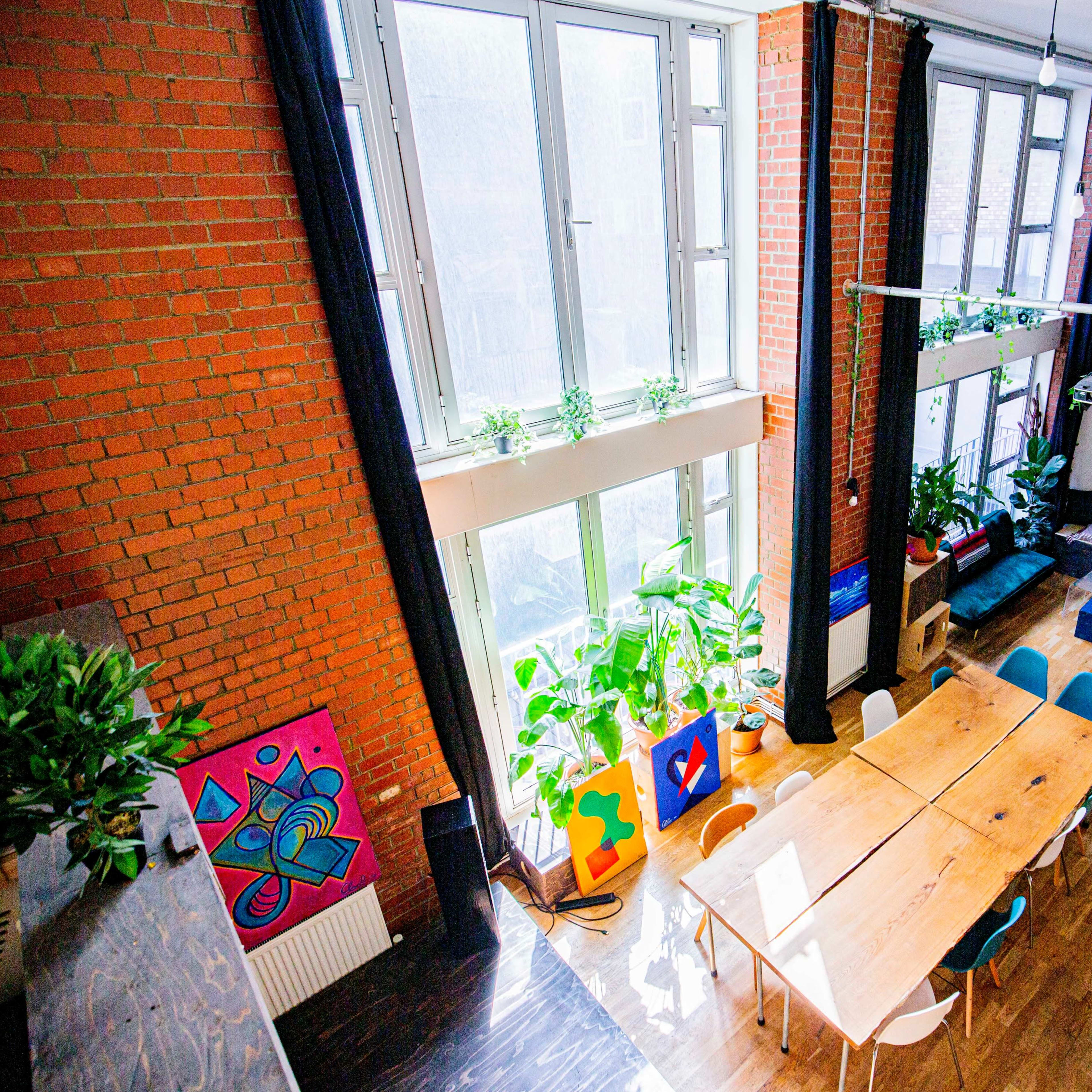 A spacious room with large windows, a long wooden table, colorful artwork on the walls, and a seating area filled with plants.