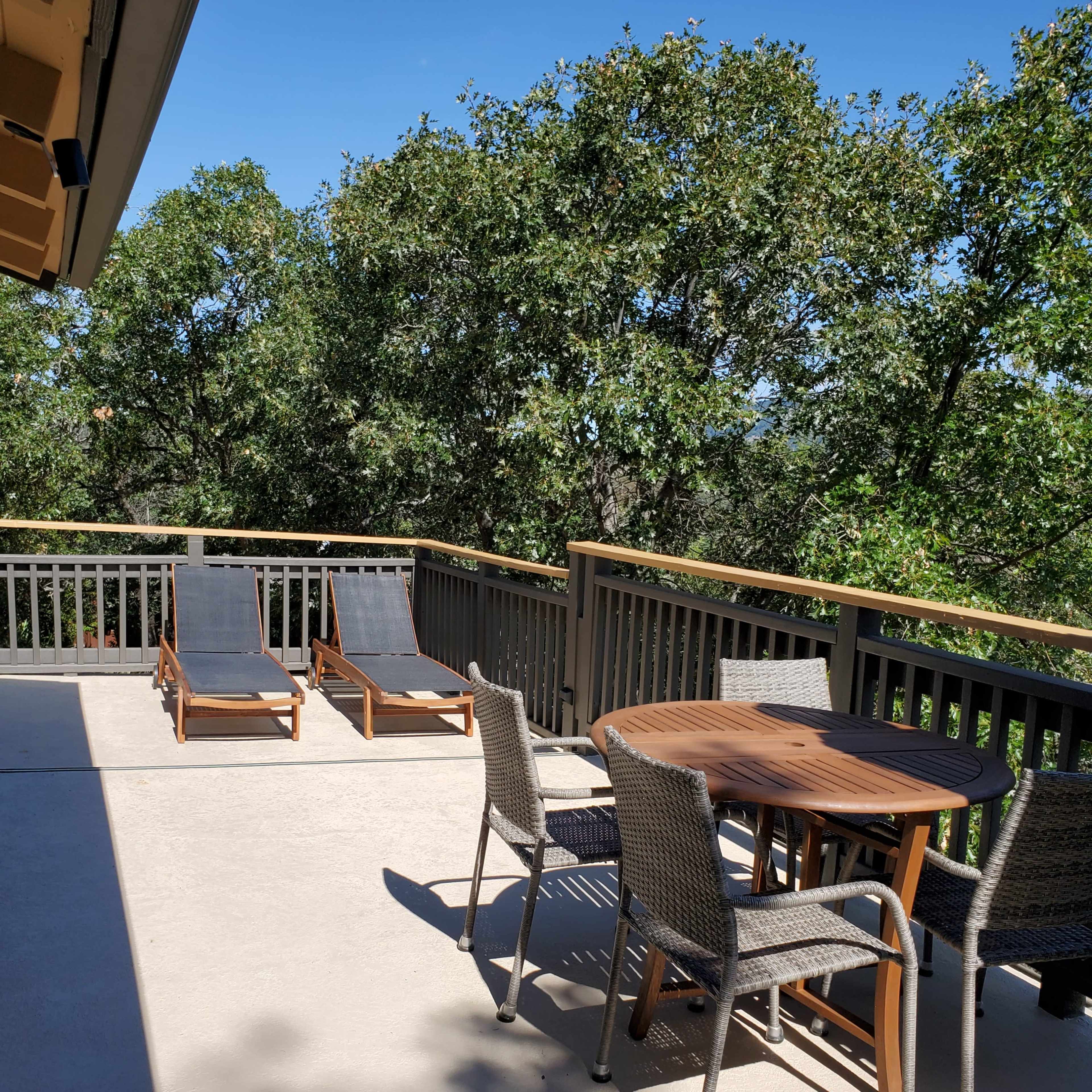 A patio area with two lounge chairs, a round table, and four chairs, surrounded by trees under a clear blue sky.