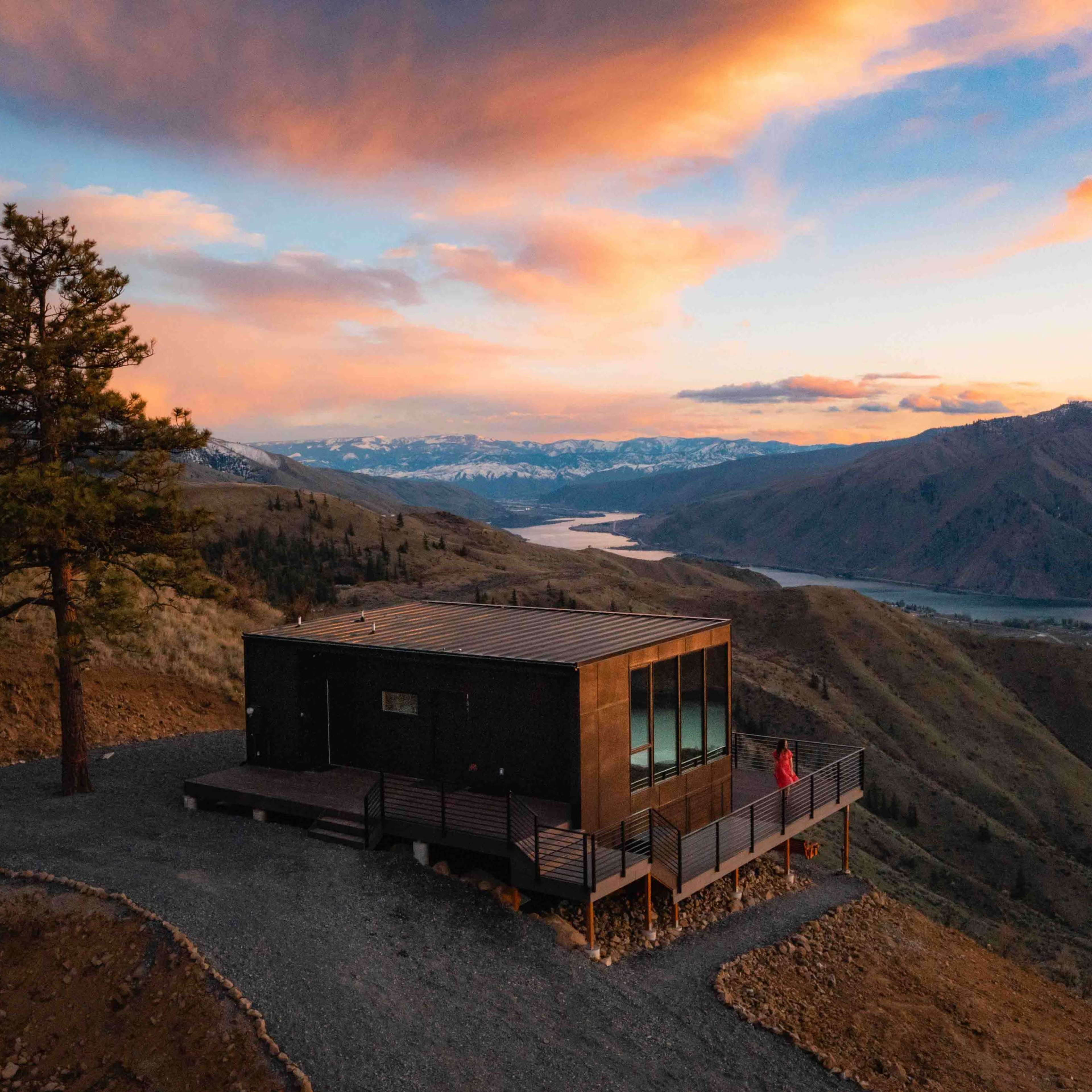 A modern cabin sits on a hillside overlooking a lake, with mountains in the background and a person standing near the edge.
