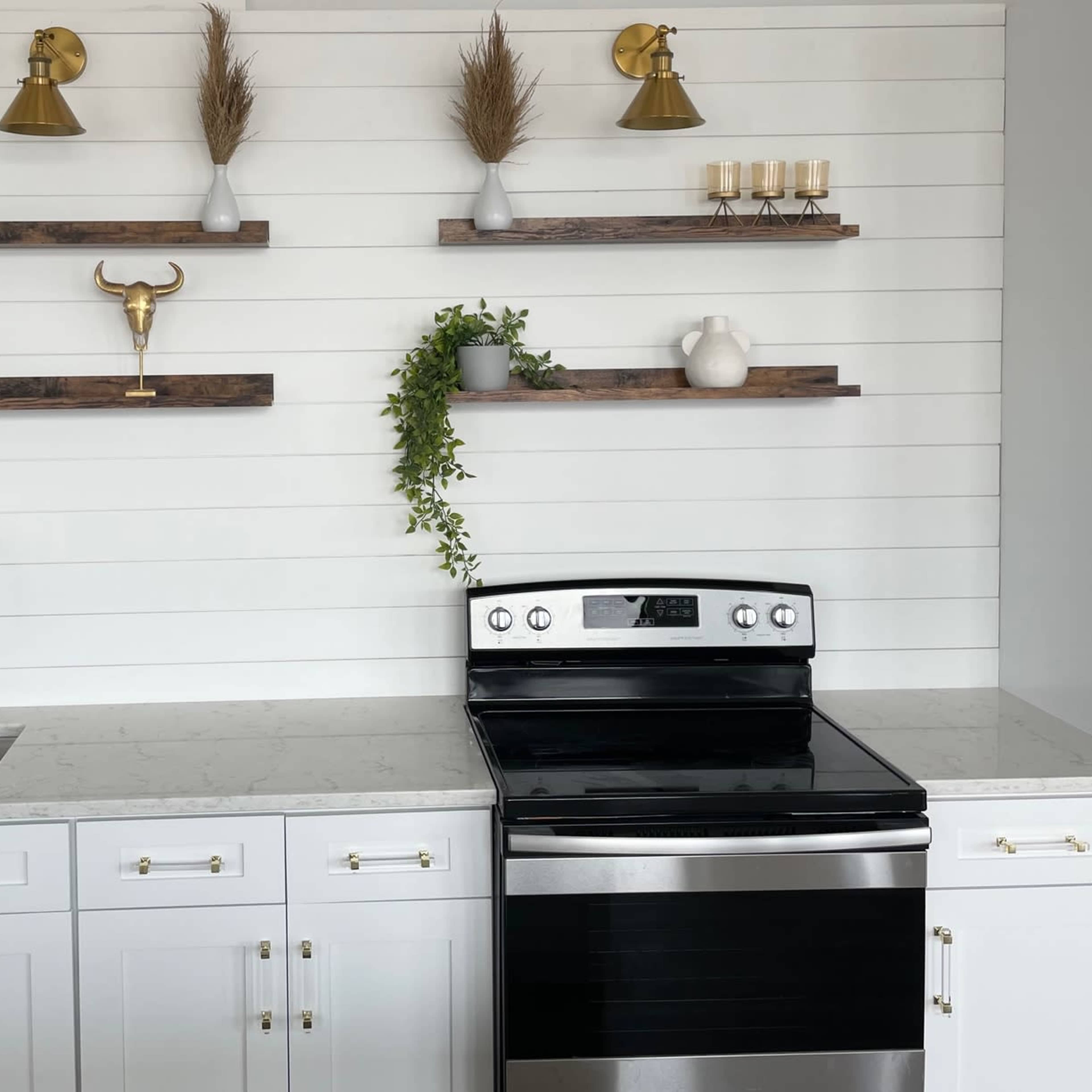 A modern kitchen features a black stove, white cabinetry, a sink with a gold faucet, and decorative shelves above with plants and accessories.