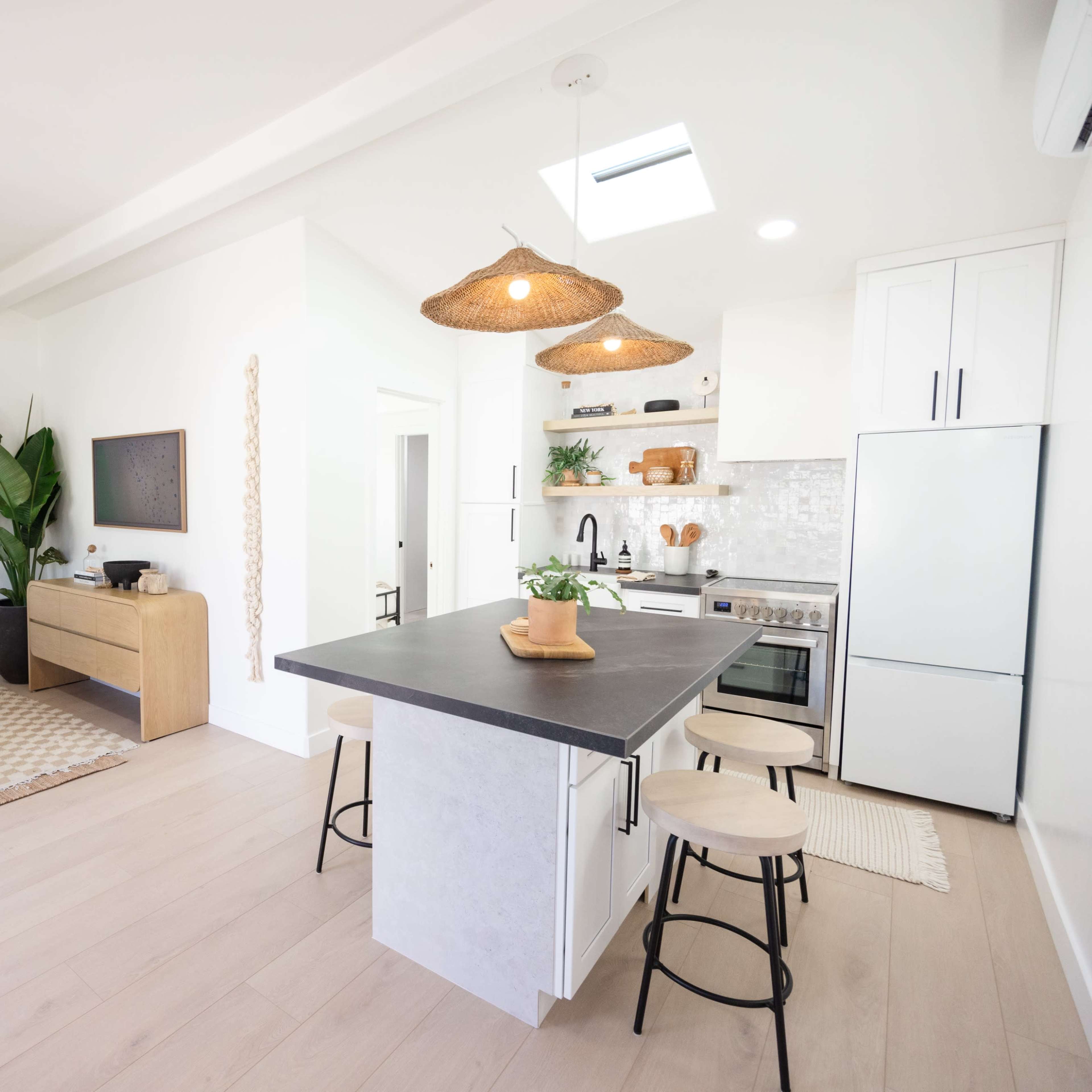 A modern kitchen and living area with light wood flooring, a kitchen island, and a large plant in the corner.
