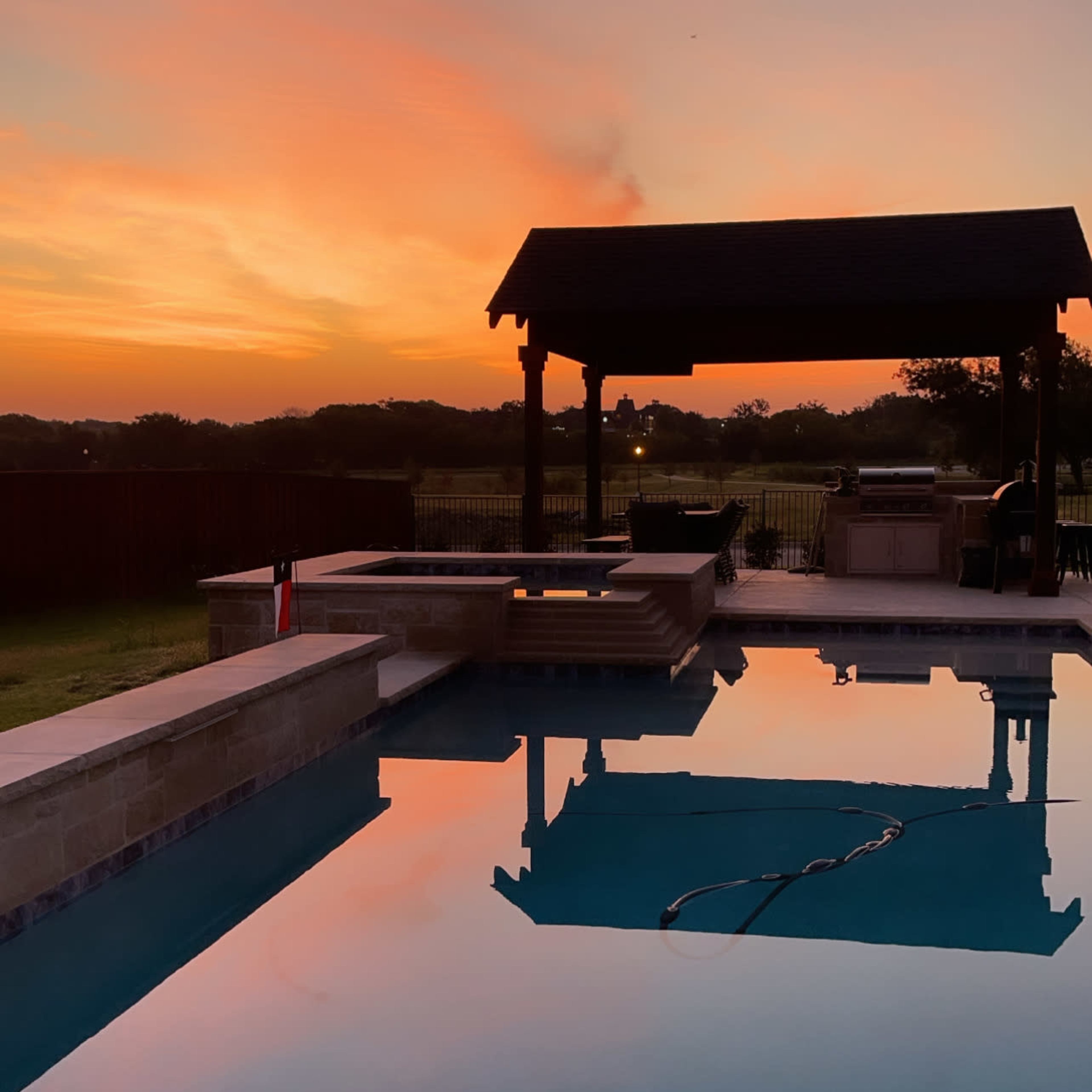 A tranquil swimming pool reflecting a colorful sunset, with a gazebo and surrounding landscape in the background.