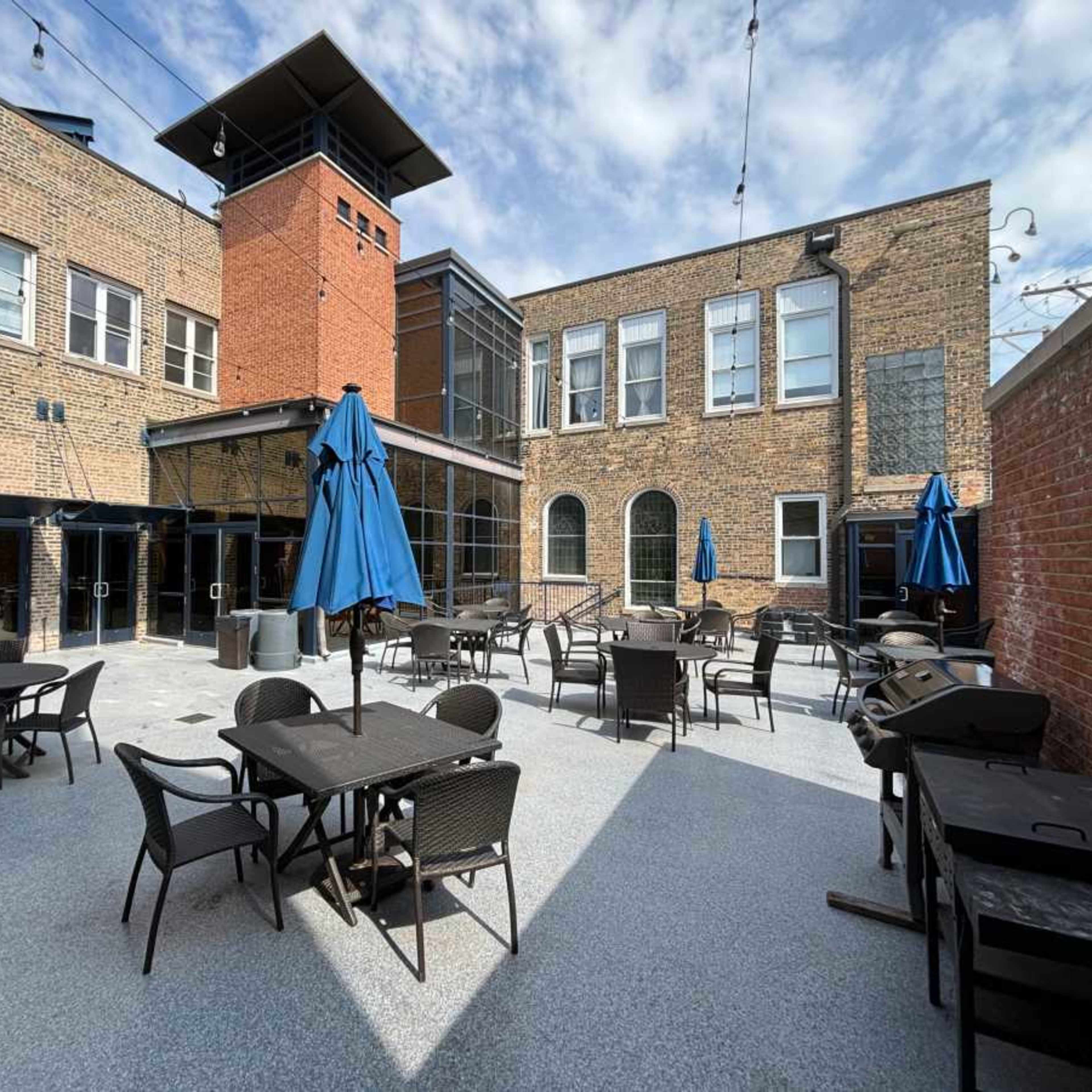 A spacious outdoor courtyard area with tables and chairs, featuring blue umbrellas and brick buildings in the background.