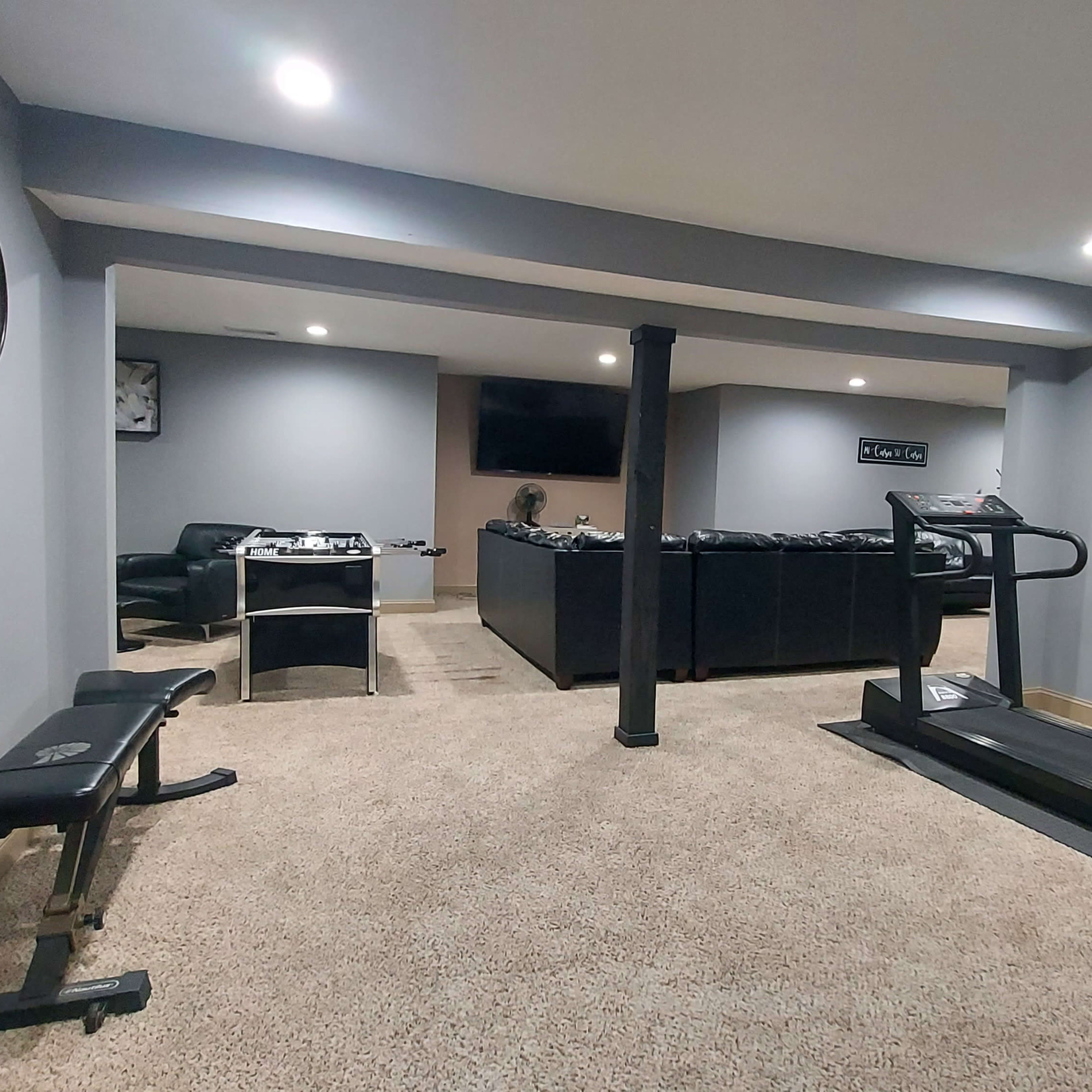 A home gym with carpeted flooring, a treadmill, a weight bench, and a desk area in a well-lit basement.