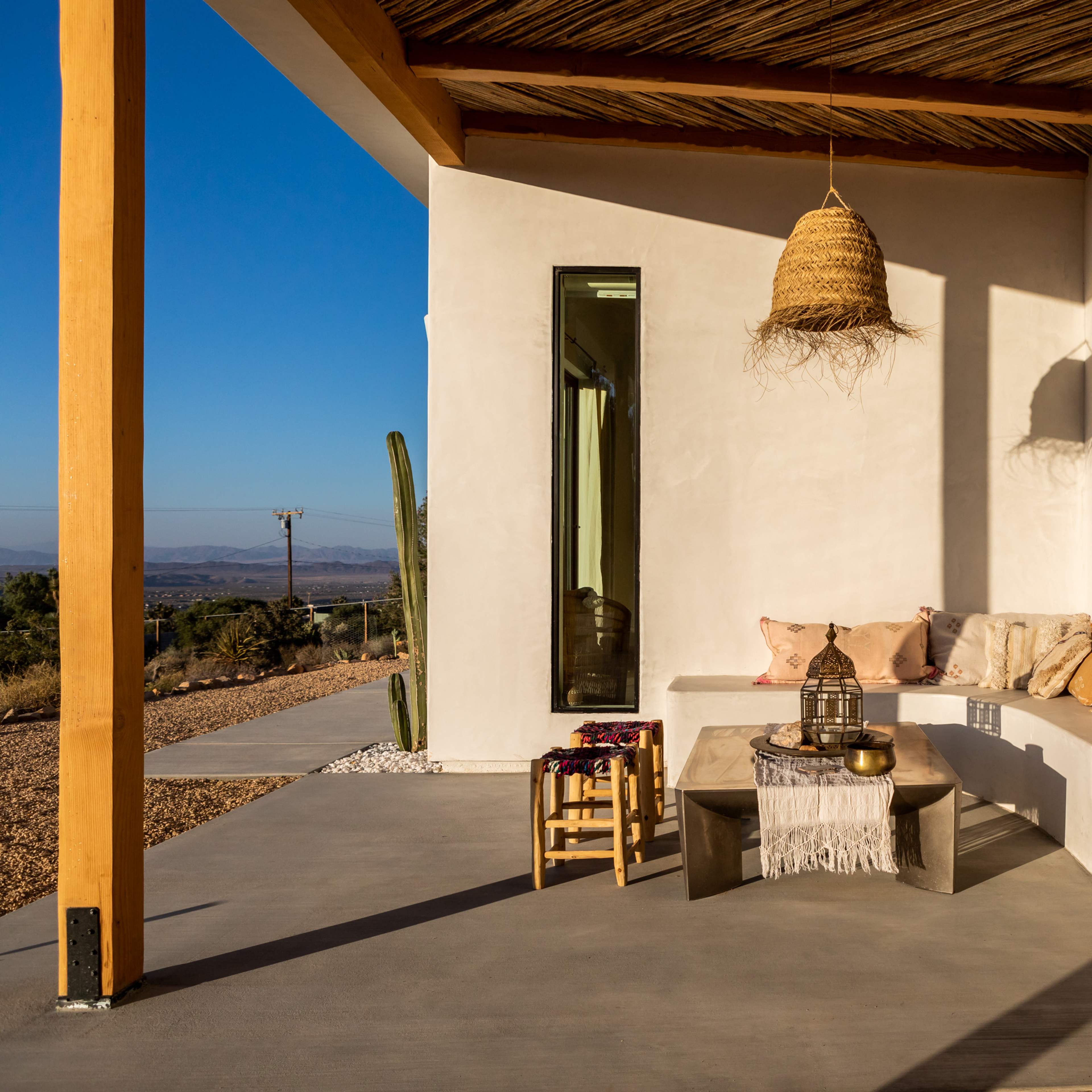 A minimalist outdoor seating area features a built-in bench with cushions, a small table, and a pendant light under a wooden overhang.
