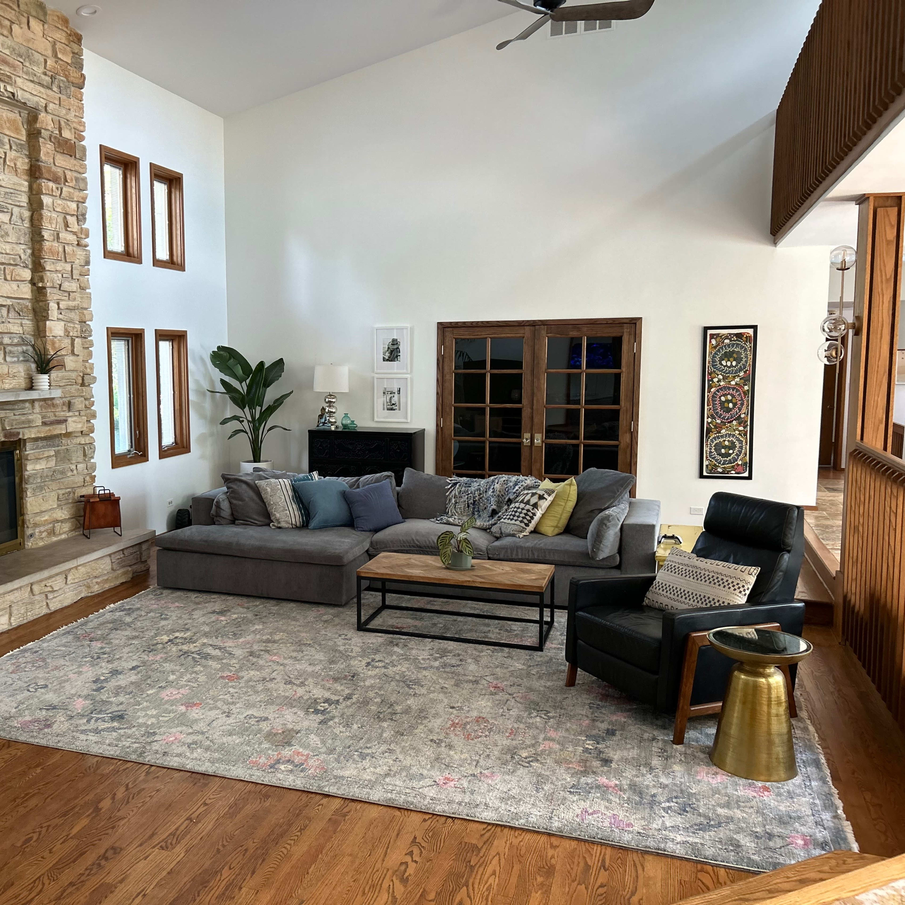 A spacious living room featuring a stone fireplace, a gray sectional sofa, a wooden coffee table, and large windows that provide natural light.