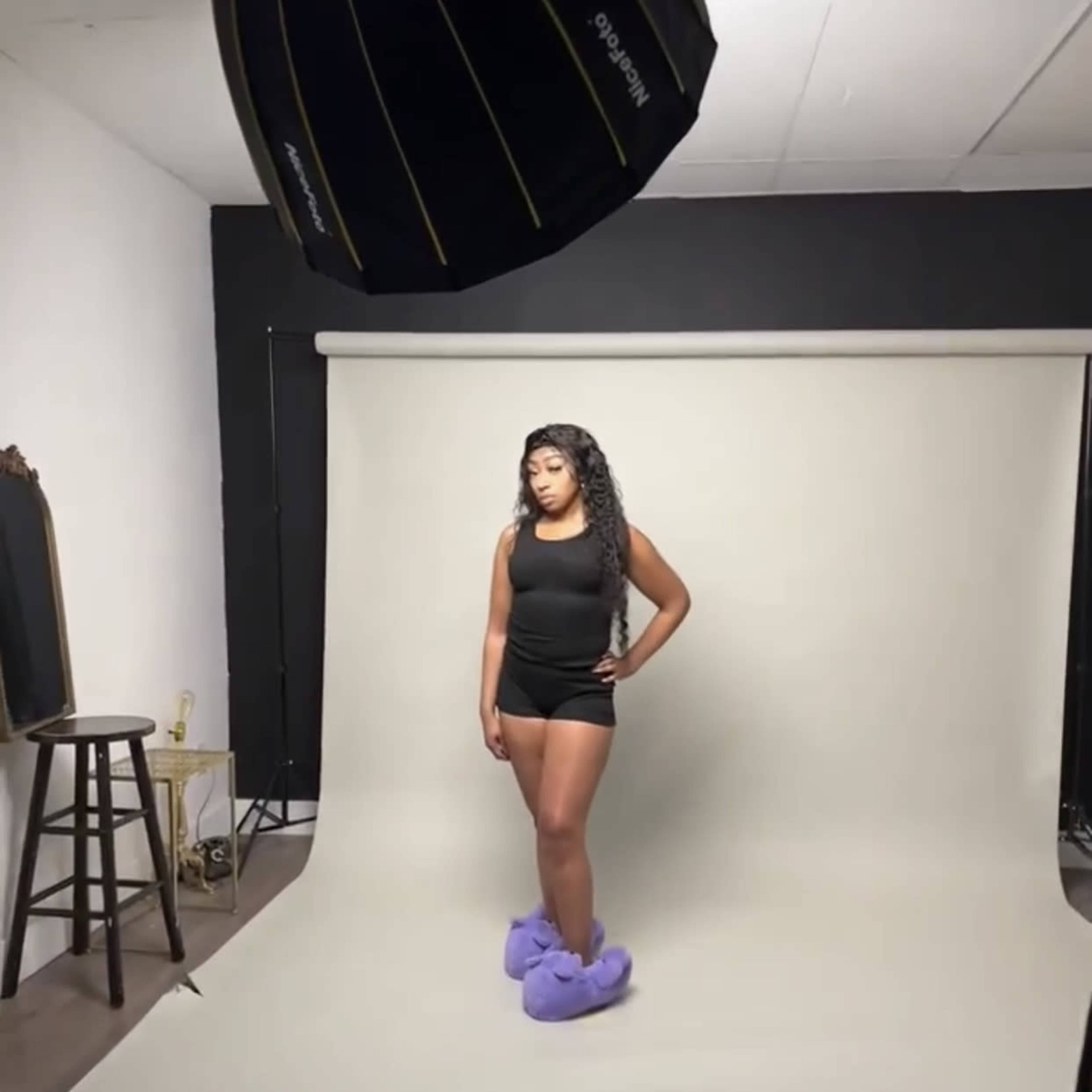 A person stands on a neutral backdrop in a photography studio, wearing a black outfit and large purple slippers.
