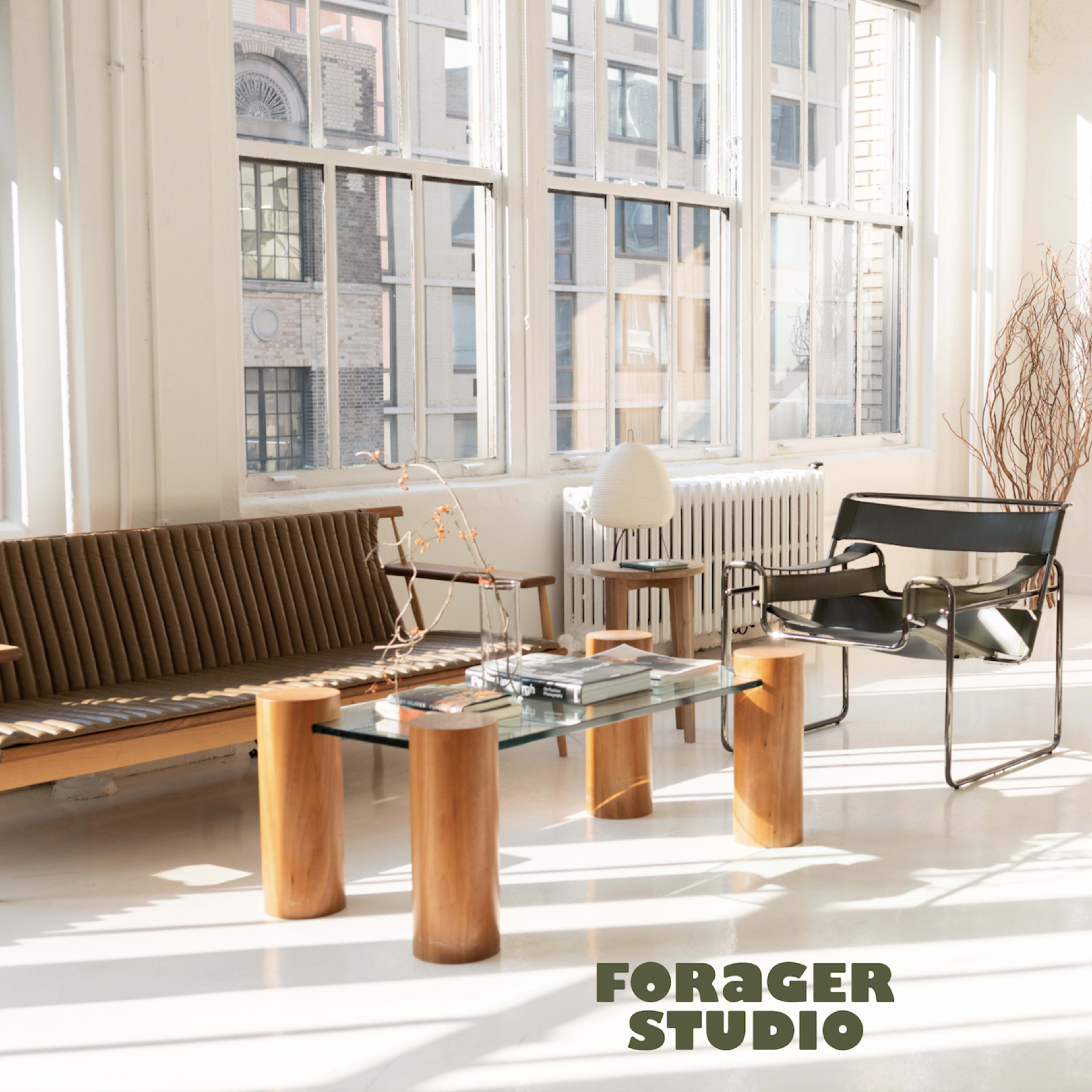 A bright studio space featuring a wooden sofa, a glass coffee table supported by wooden columns, and a minimalist black chair, with sunlight streaming through large windows.