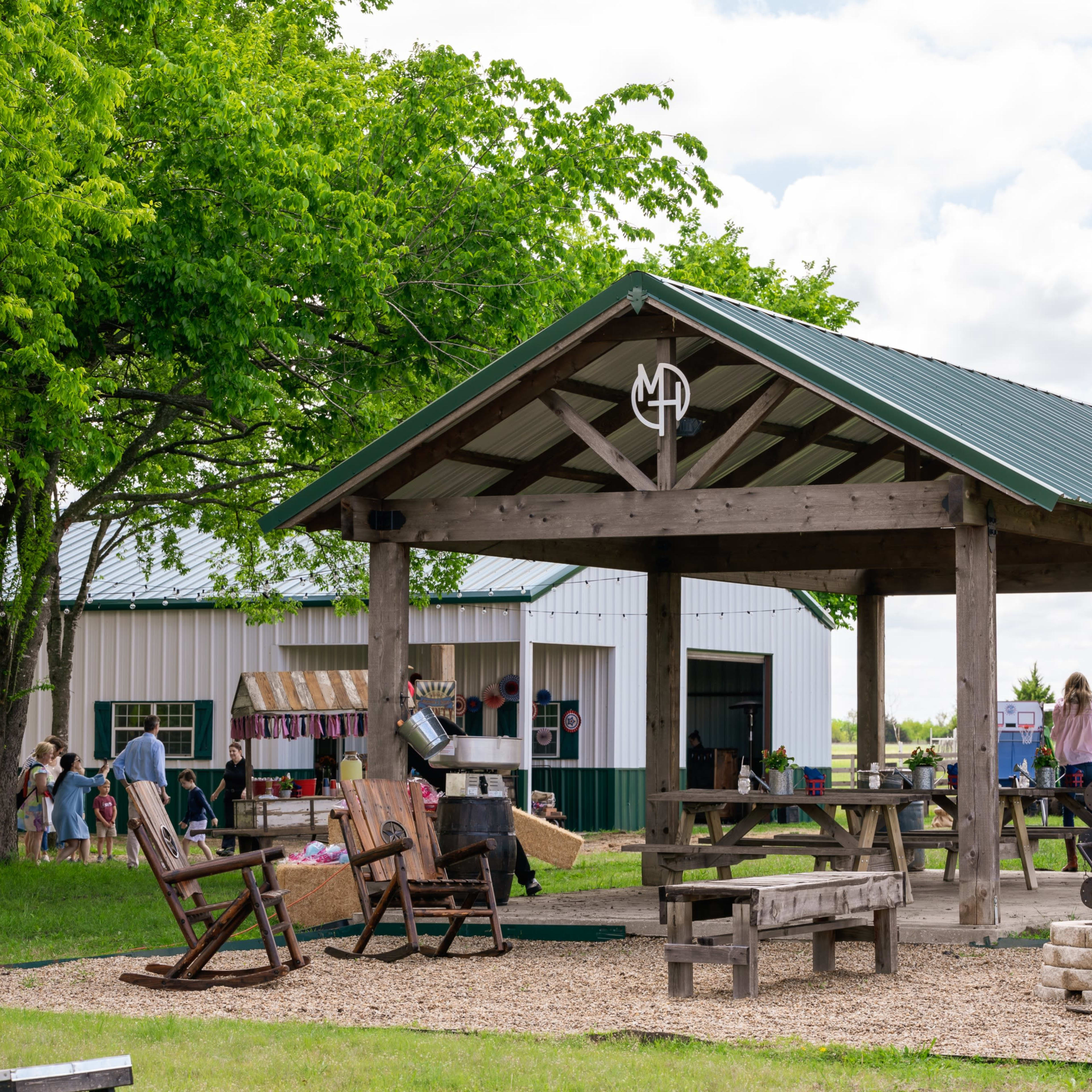 A wooden pavilion with a metal roof sits in a green space surrounded by trees, with several people engaging in activities nearby.