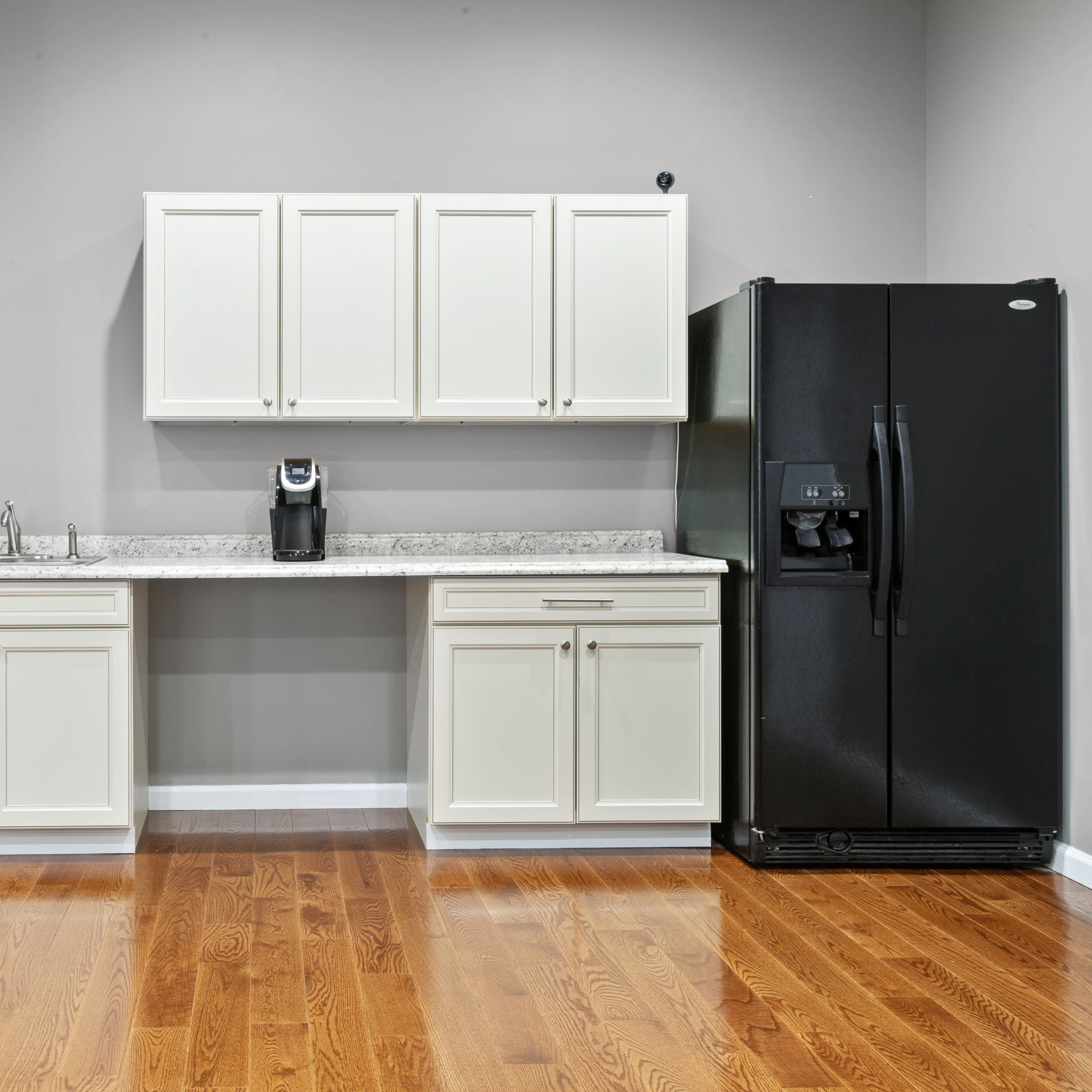 A kitchen area with white cabinetry, a countertop, a coffee maker, and a black refrigerator against a gray wall.