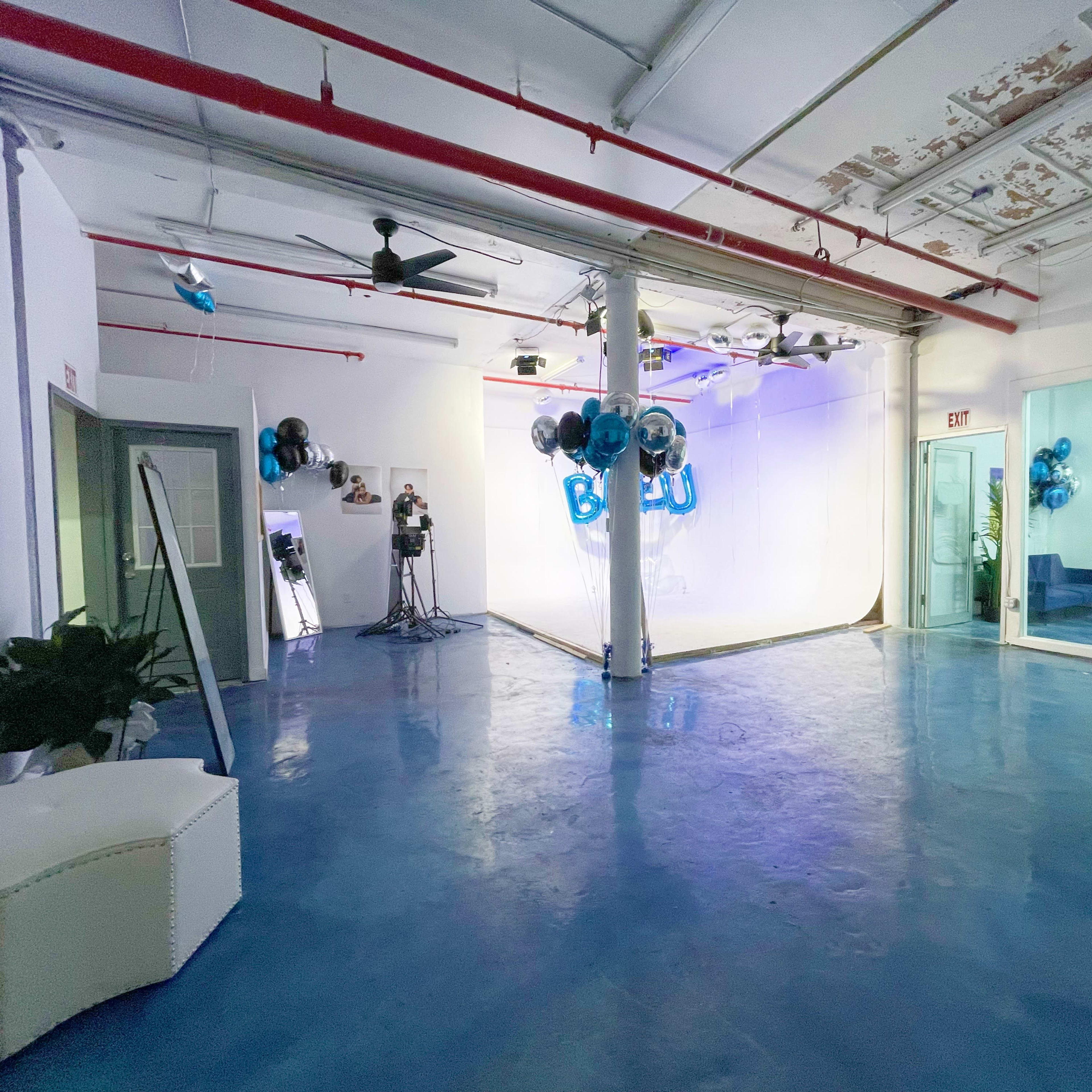 An empty event space with a blue floor, white walls, and decorations of blue and silver balloons, along with a glass-walled area featuring chairs and a table.