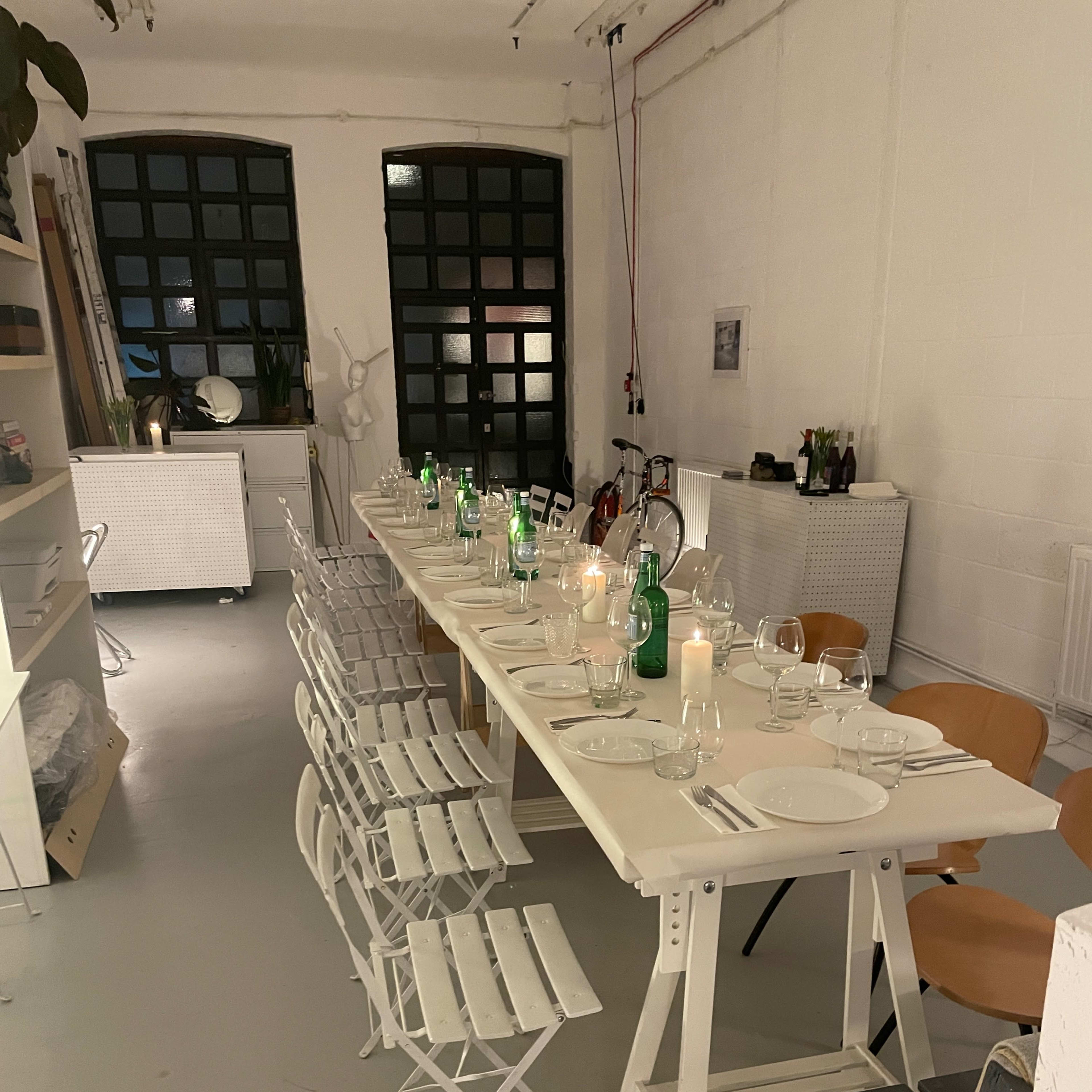 A long table is set for dinner, surrounded by various chairs, with candles and green bottles in a well-lit, minimalist room.