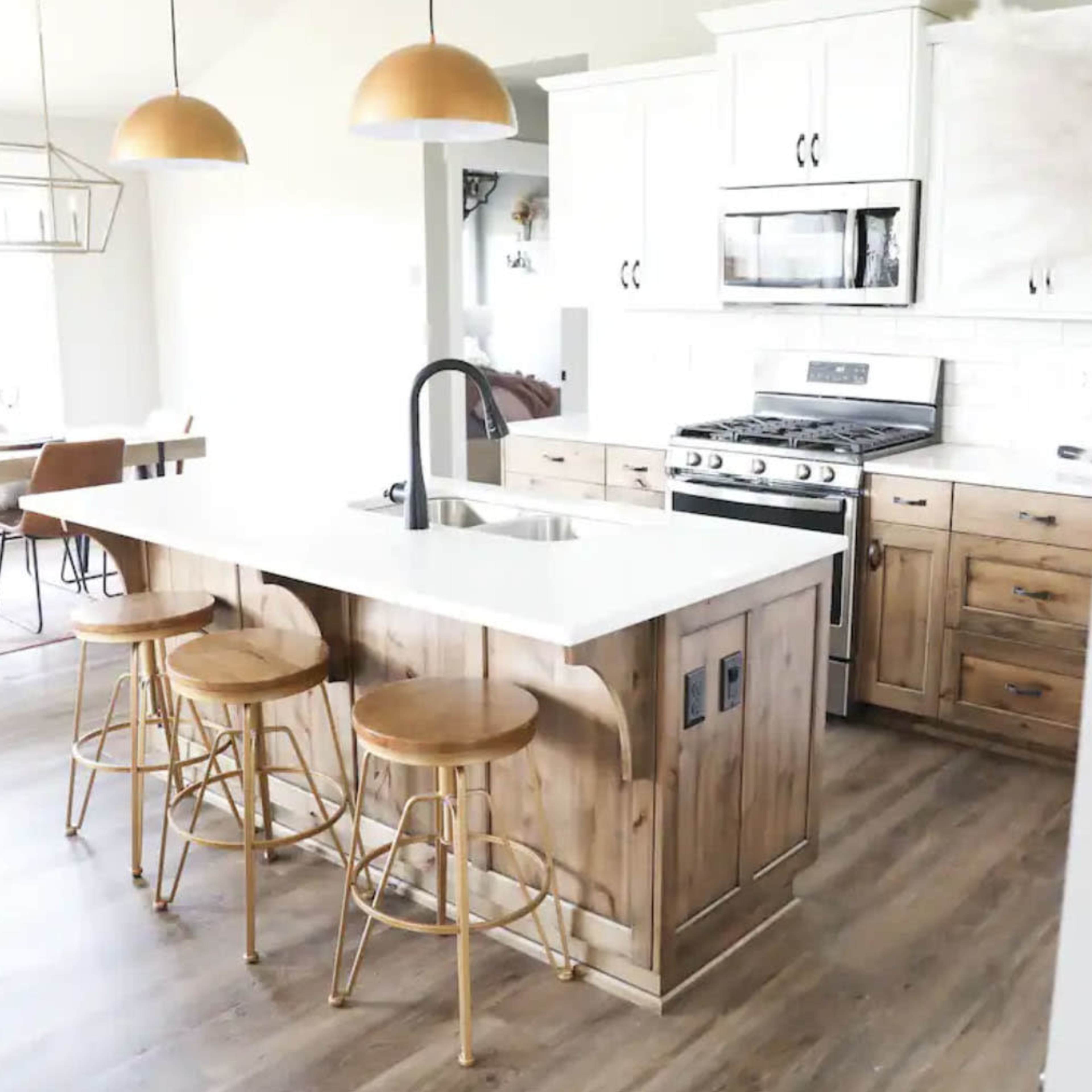 A modern kitchen features a central island with three wooden stools, equipped with a sink and surrounded by light-colored cabinets, a stainless steel stove, and large windows.