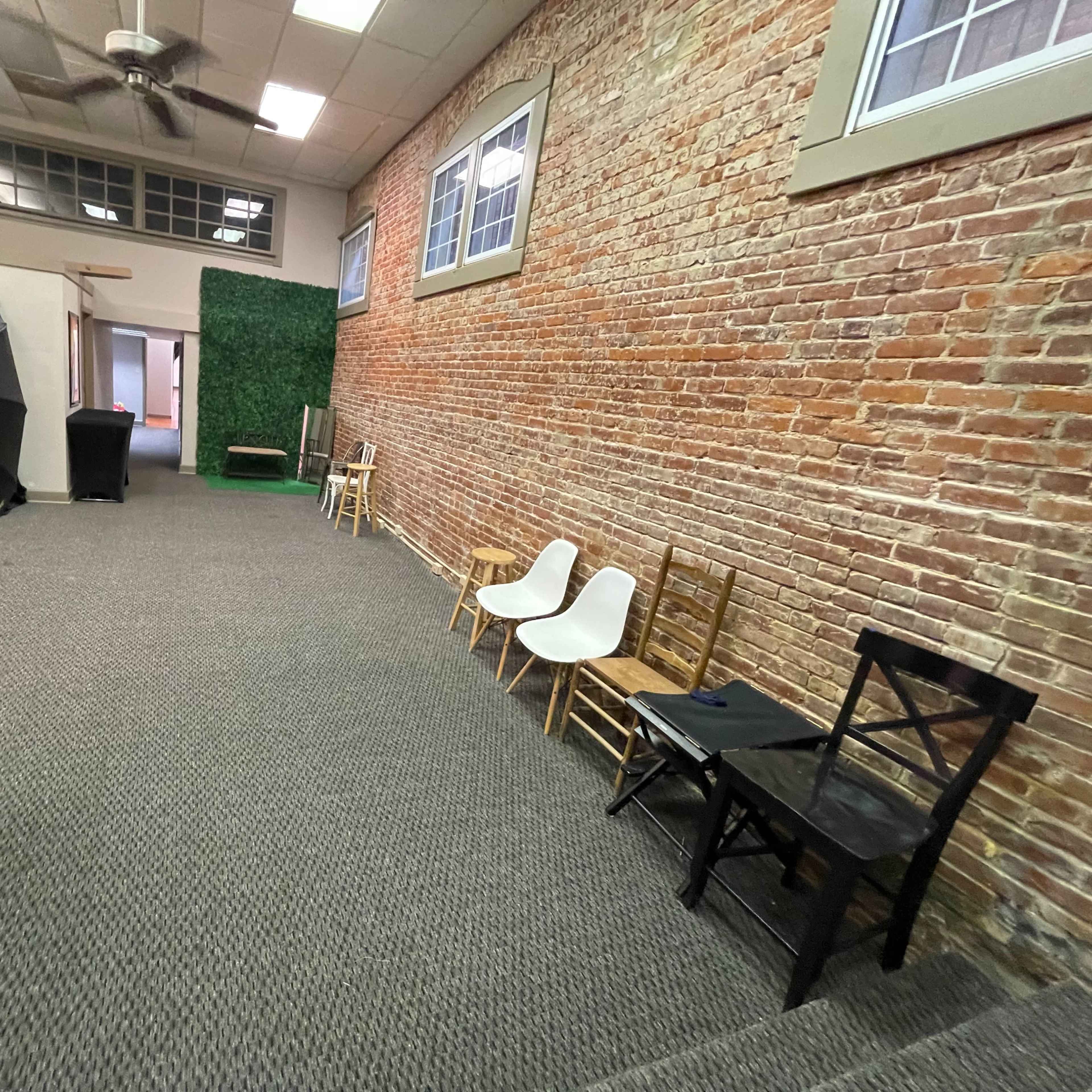 An interior space with a brick wall, a carpeted floor, and several chairs arranged along one side, alongside a large black umbrella reflector.