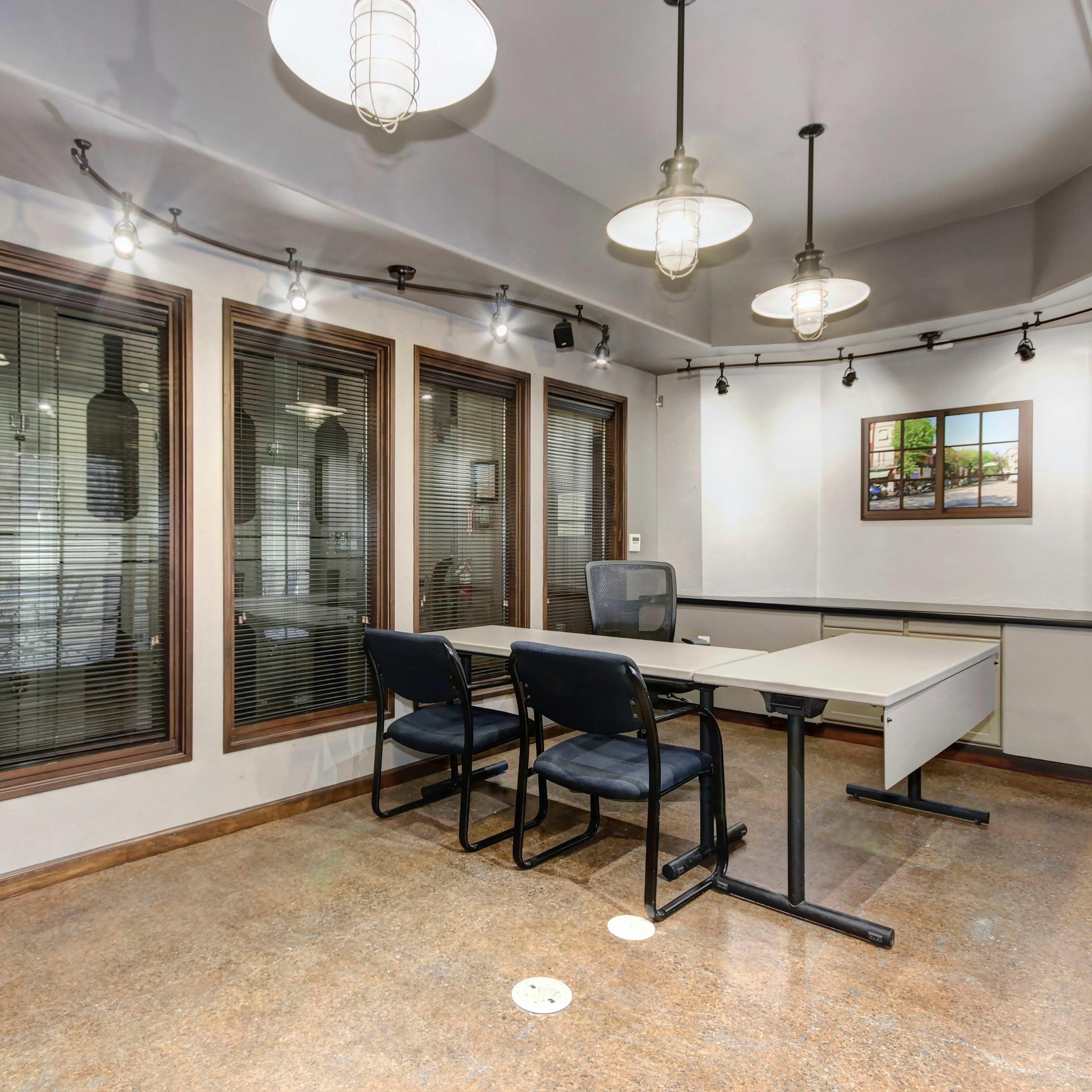 A modern office space with a central desk and two chairs, surrounded by large windows framed by dark blinds.