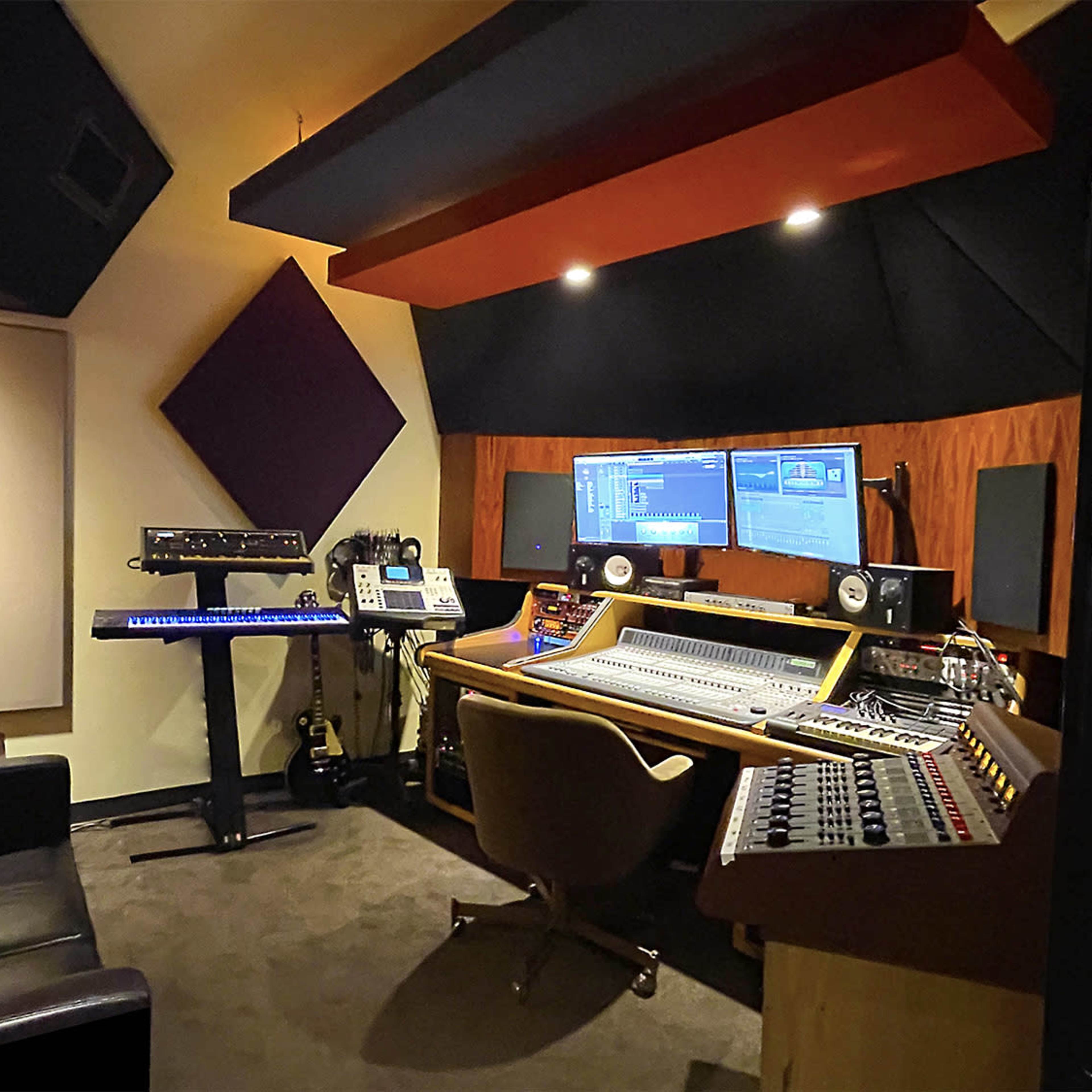 A music studio with two monitors, a mixing console, and various audio equipment, including a keyboard and a lamp.
